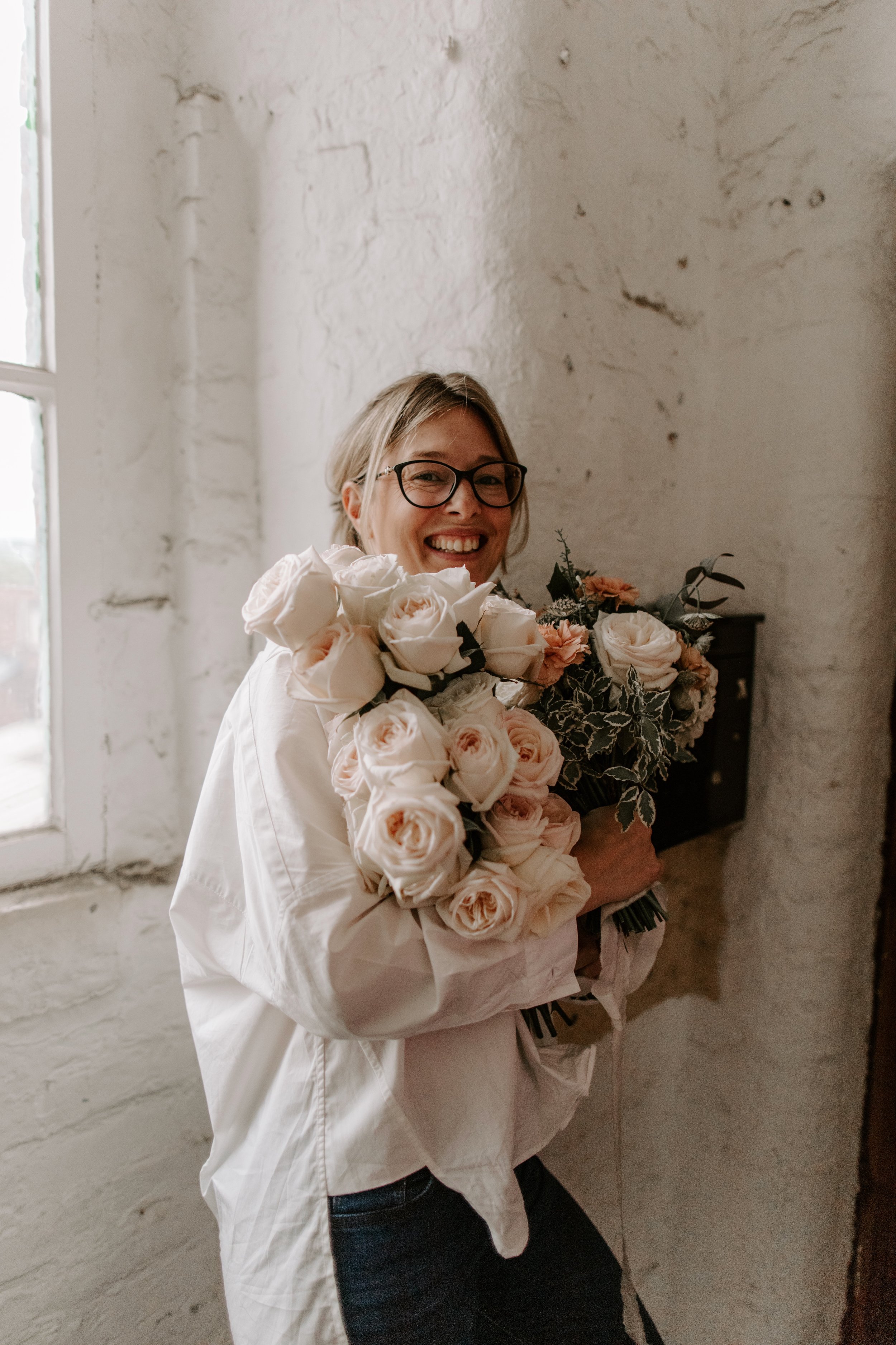 Smiling person holding bouquets of white and peach roses in a rustic indoor setting.