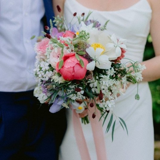 Bride holding colorful wedding bouquet with various flowers, standing next to groom in suit.