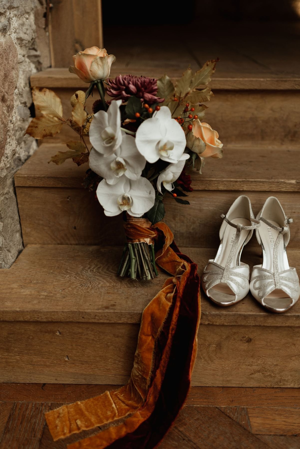 A bouquet of white orchids and peach roses with autumn leaves on wooden steps, next to a pair of white bridal shoes with rhinestone details.