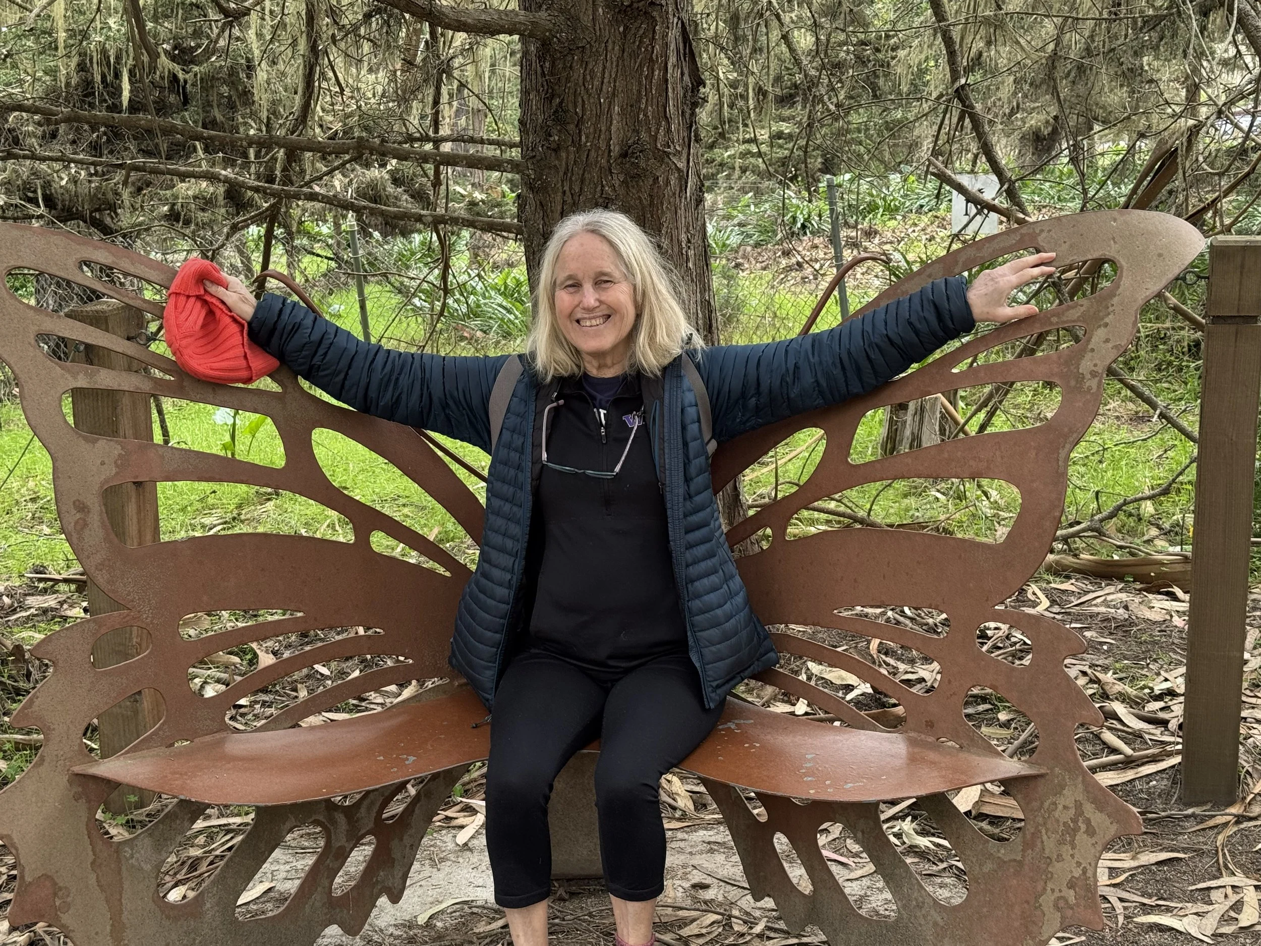 A woman with blonde hair sitting on a butterfly-shaped metal bench in a wooded outdoor area, smiling with arms outstretched on the bench wings, wearing a black jacket and black pants.