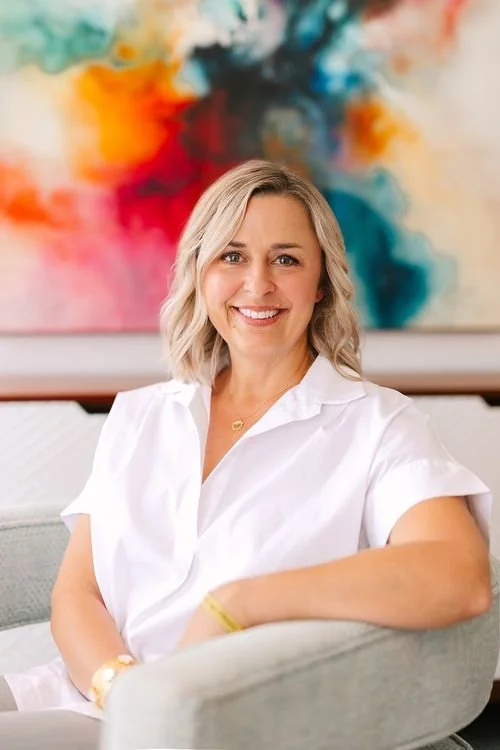 A smiling woman with shoulder-length blonde hair wearing a white shirt, sitting on a light-colored couch in front of an abstract colorful painting.