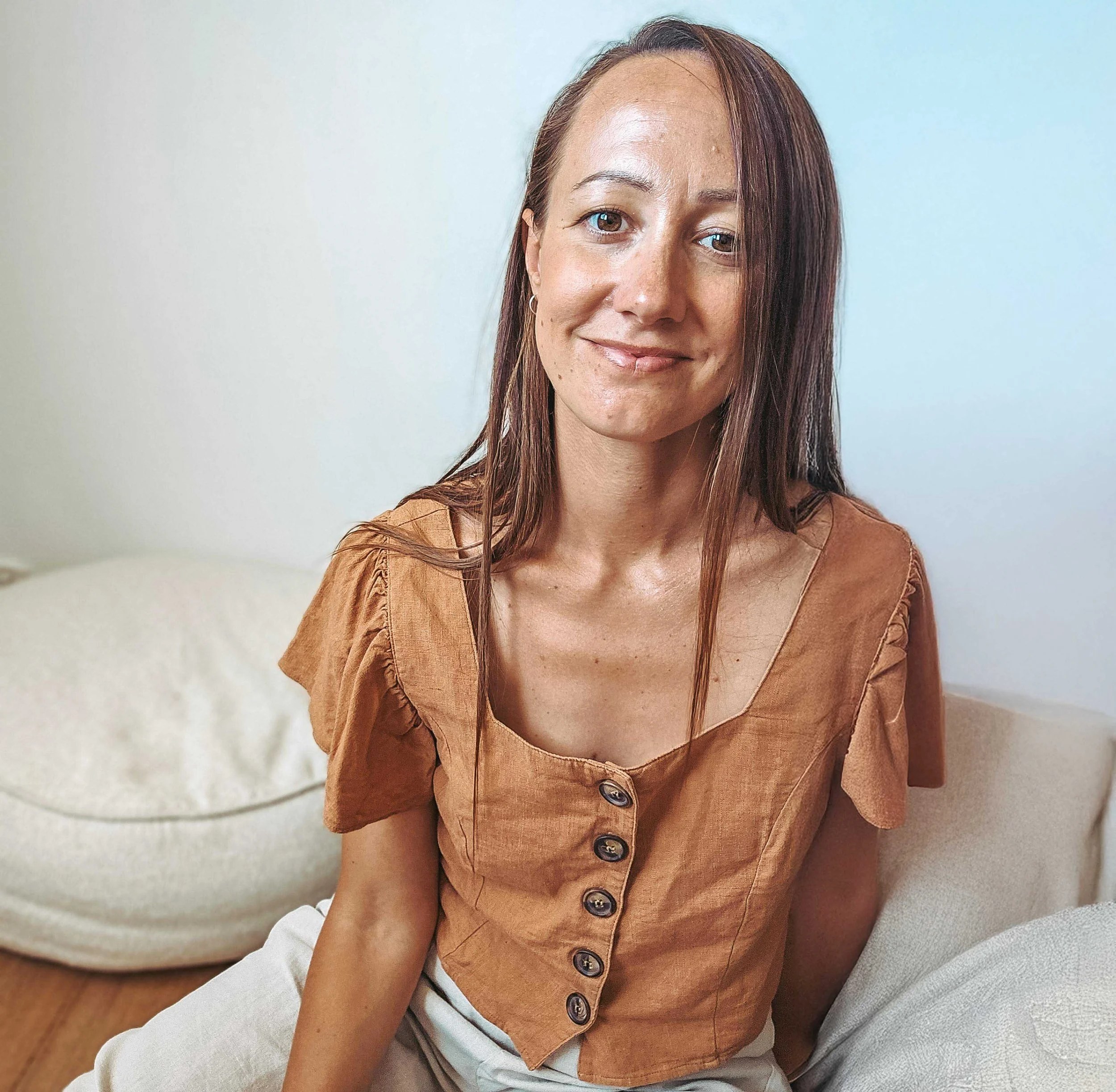 A woman with light skin, long brown hair, and a gentle smile sitting on a beige couch in a light-colored room.