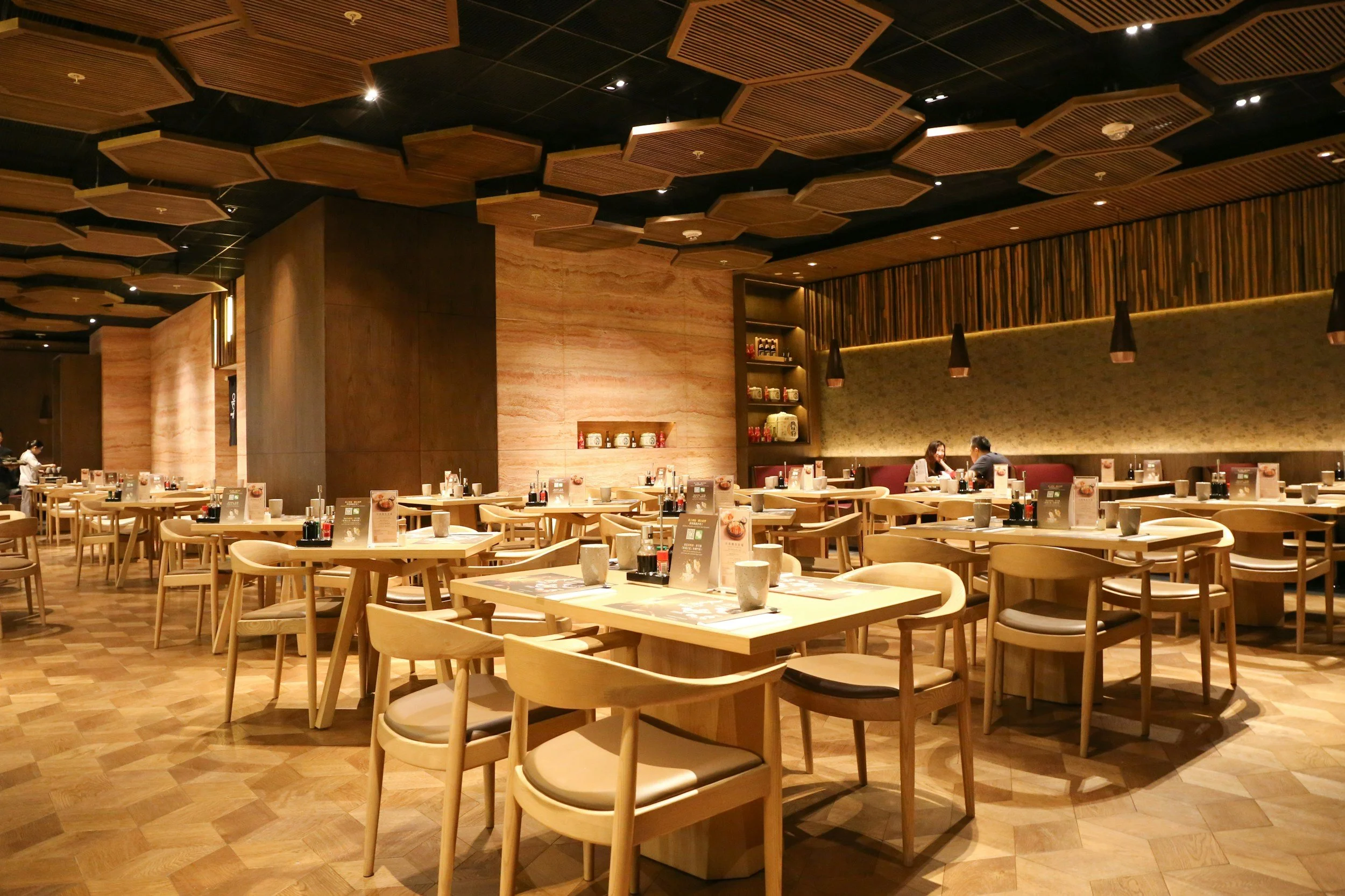 wide angle shot of an almost empty restaurant with wood effect parquet flooring, light coloured informal chairs and tables and modern architectural geometric wood ceiling