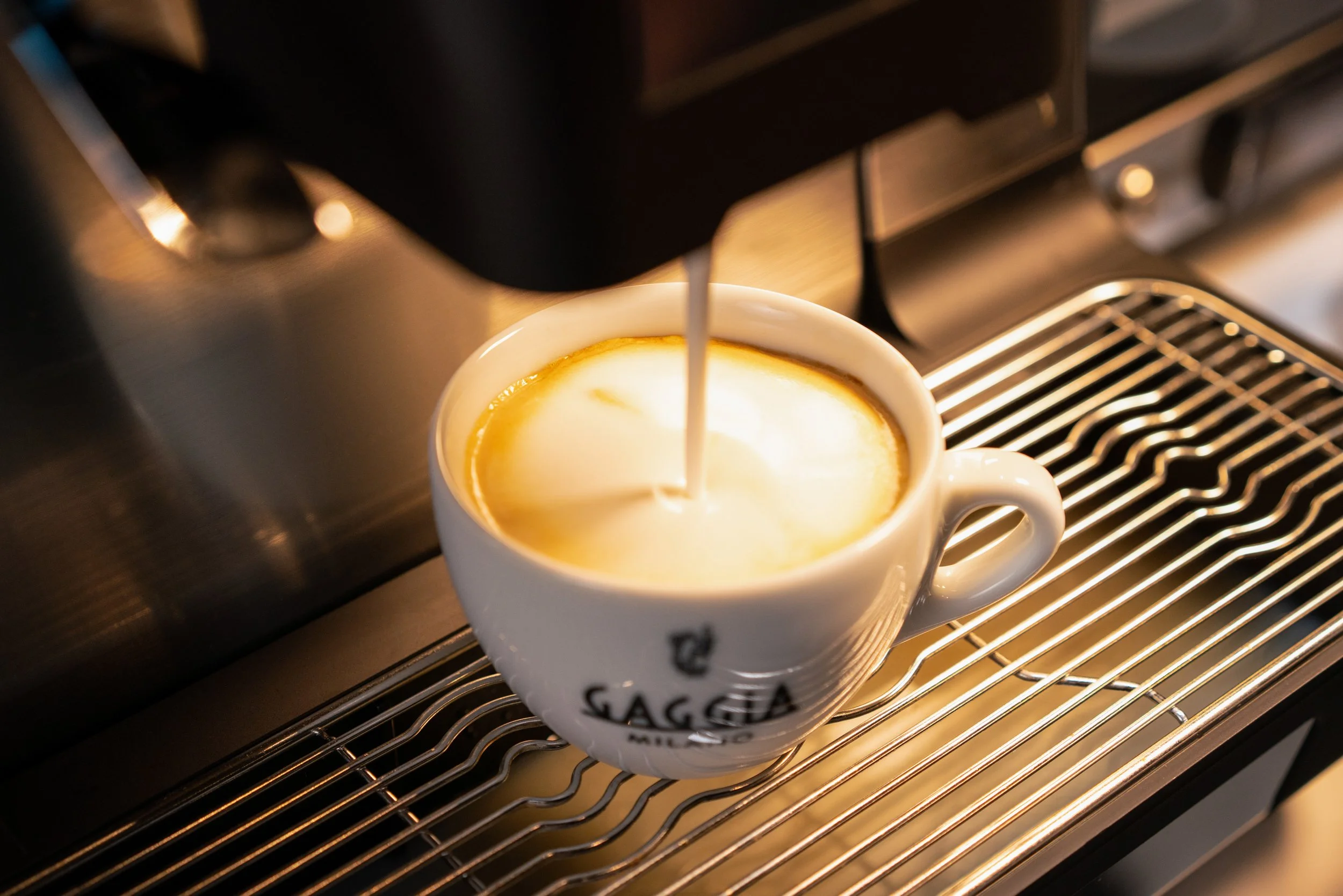 an artistic photograph of a white coffee cup diplaying Gaggia branding sat on a coffee machine being filled with latte