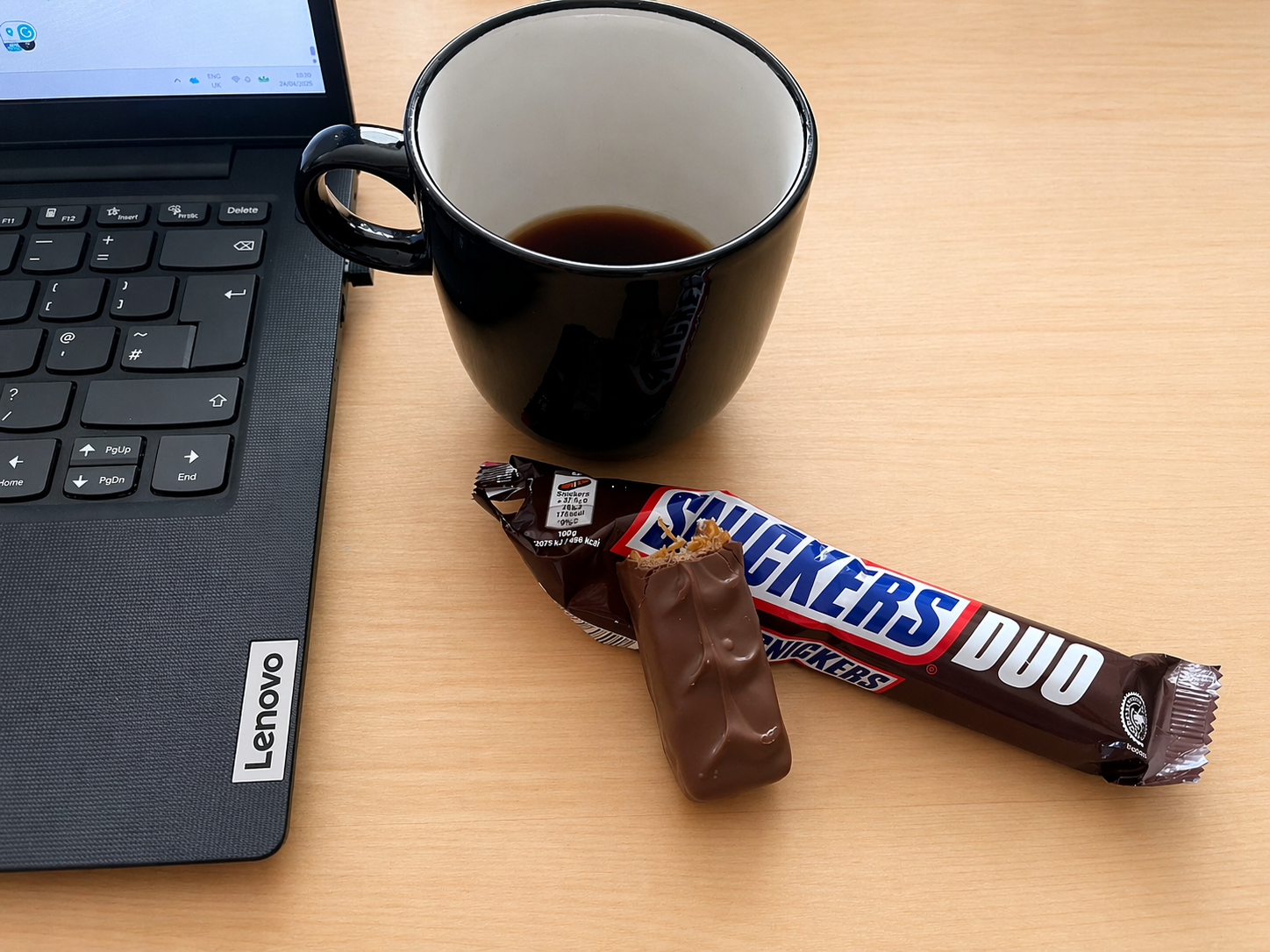 clean modern photo of a half drunk black coffee in a stylish black mug sat on a light wooden desk alongside an open laptop, along with a half eaten bar from an open snickers duo pack