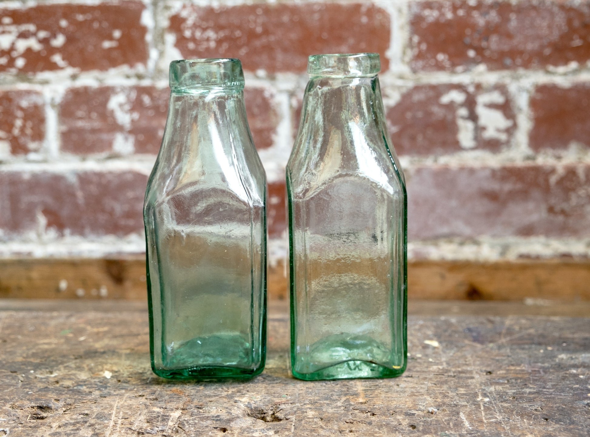 a photograph of 2 antique vintage square glass traditional milk bottles stood on a concrete floor, in front of a rustic red brick wall