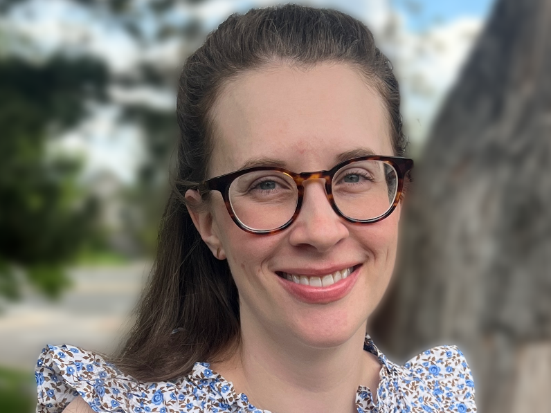 A woman with long brown hair, glasses, and a floral-patterned top smiling outdoors, with blurred trees and sky in the background.
