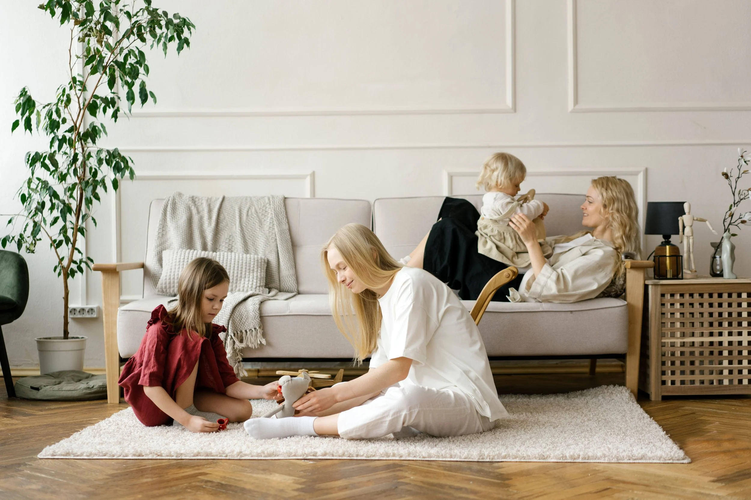 Two moms and their children playing with toys on a beige carpet in a living room, with a white couch and decorative plants in the background.