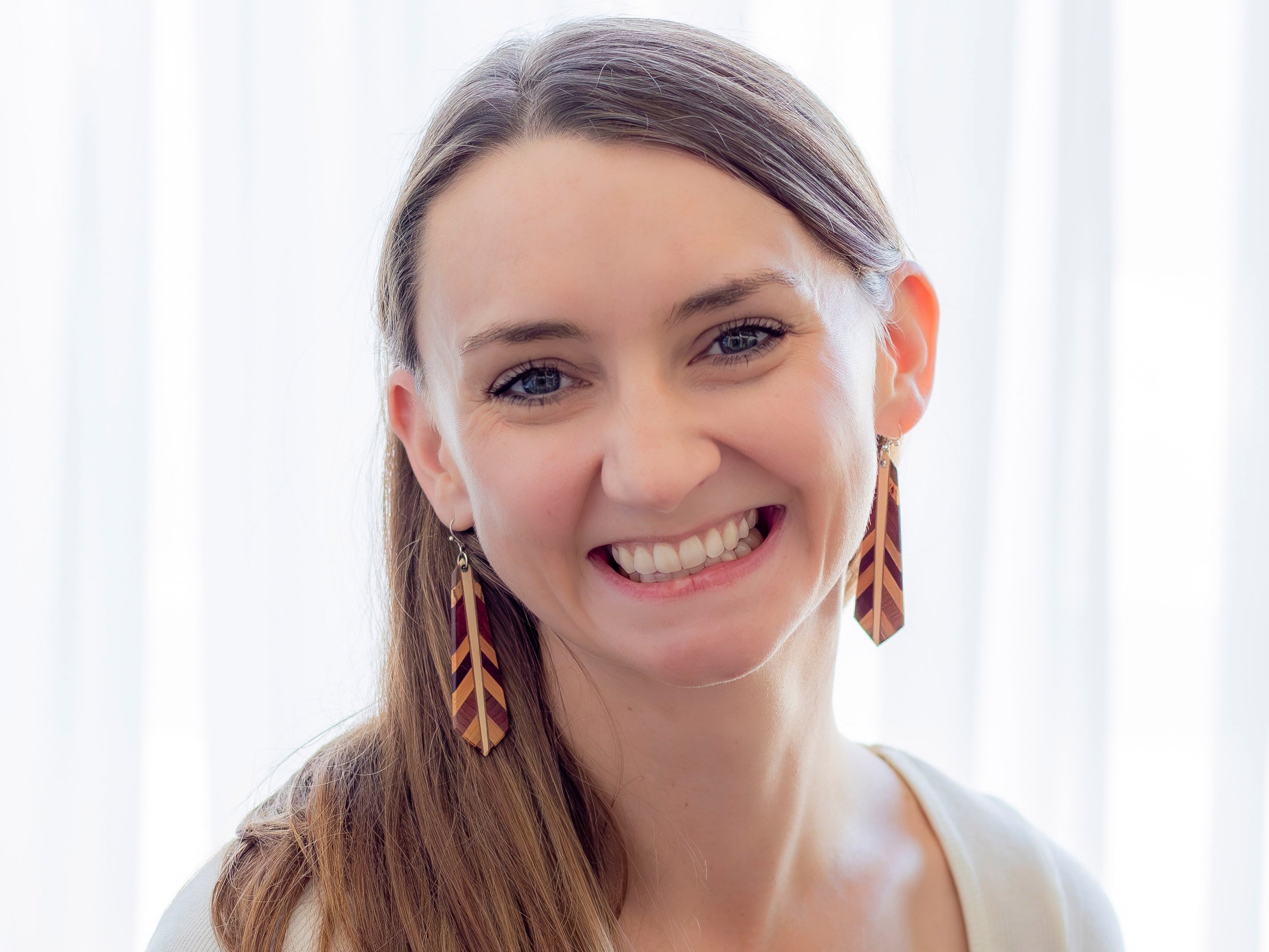 Close-up of a young woman's face with a big smile, showing her teeth, wearing colorful feather earrings, with light brown hair and a white top.