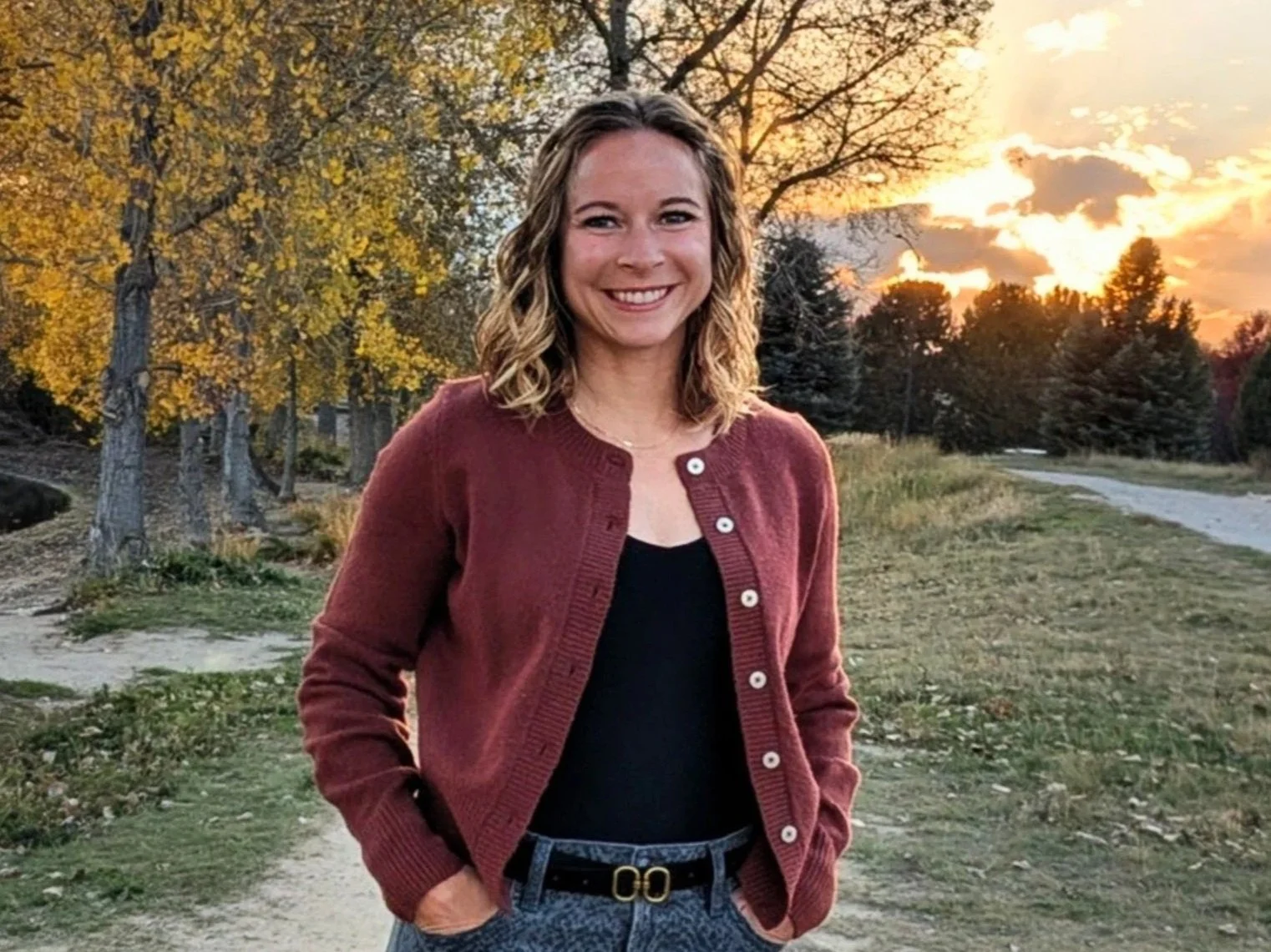 A young woman with curly blonde hair standing outdoors on a dirt path during sunset, with trees and orange sky in the background.
