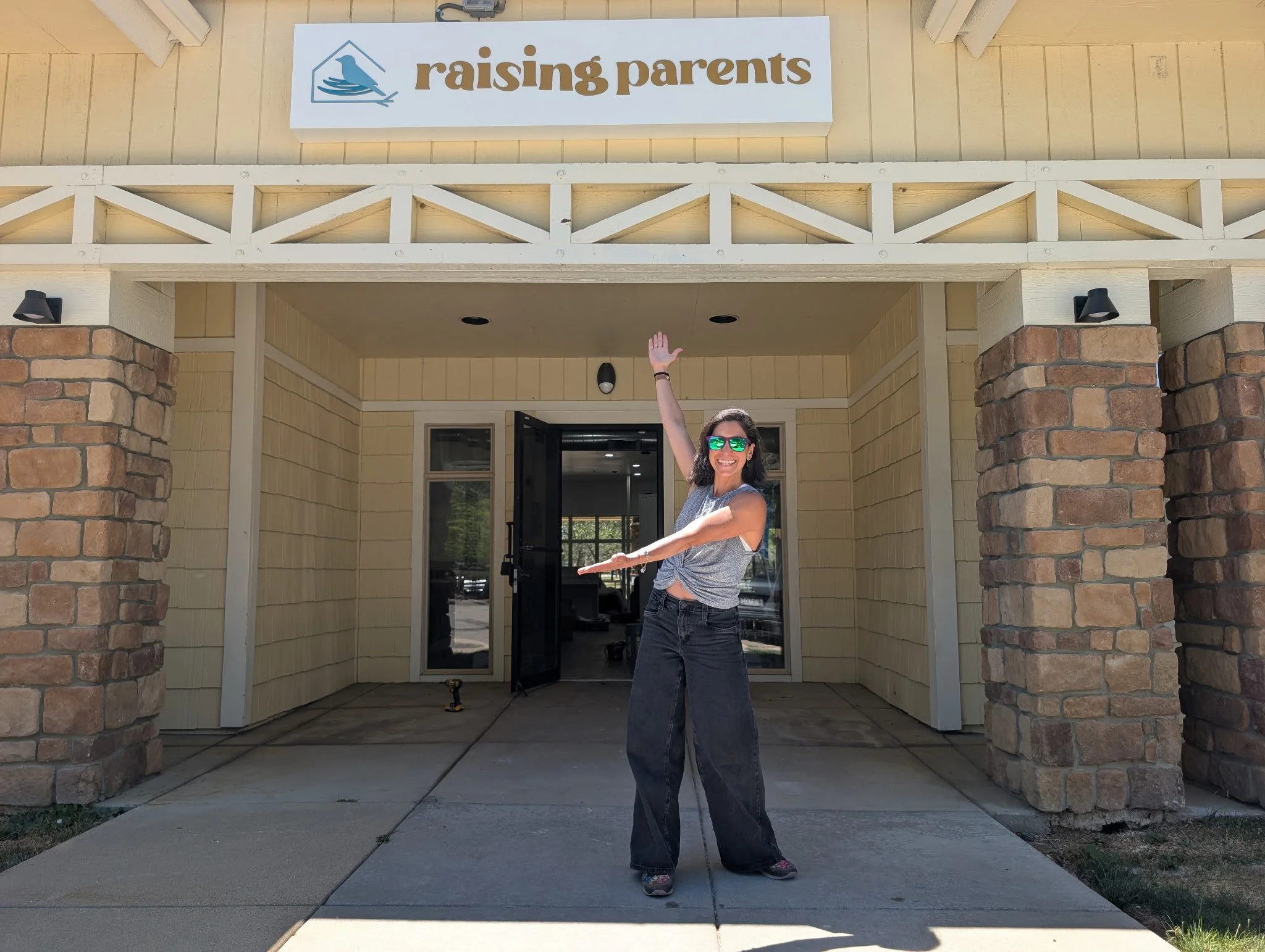 Maha wearing sunglasses and casual clothing, smiling and raising one arm, standing in front of a building with a sign that reads "raising parents".