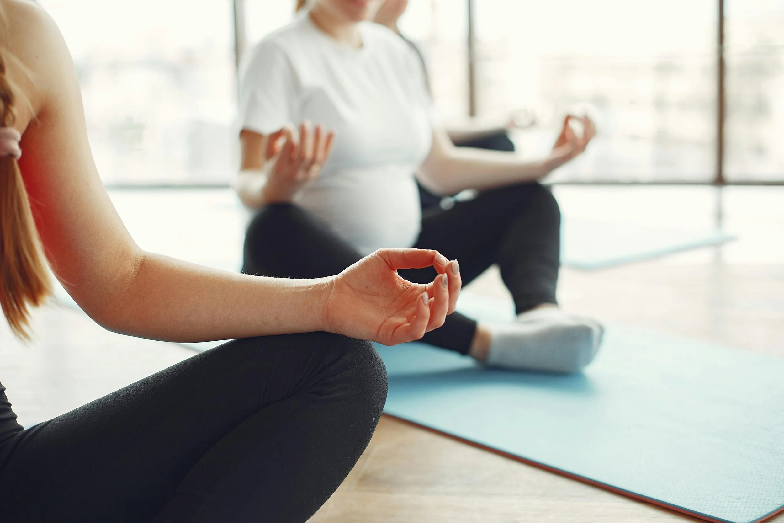 Close-up of a pregnant woman practicing yoga or meditation, sitting cross-legged on a blue yoga mat, with hands in a mudra position, indoors with large windows in the background.