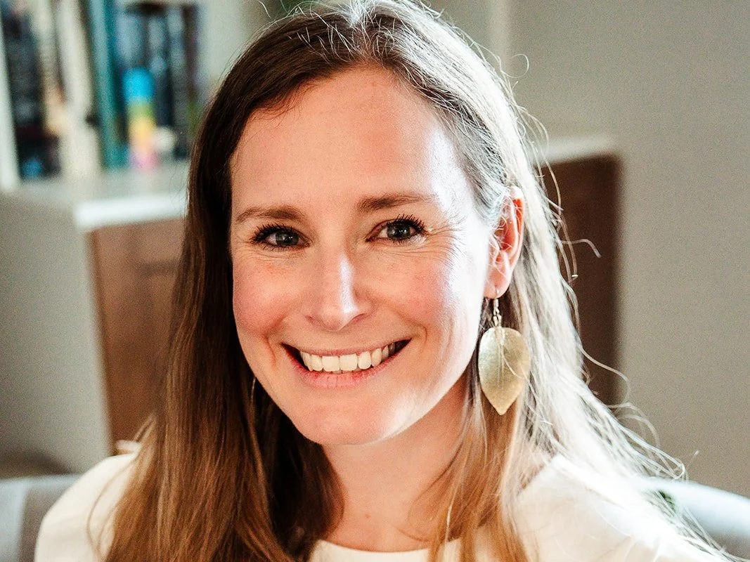 A smiling woman with long brown hair and large leaf-shaped earrings, indoors with bookshelves in the background.