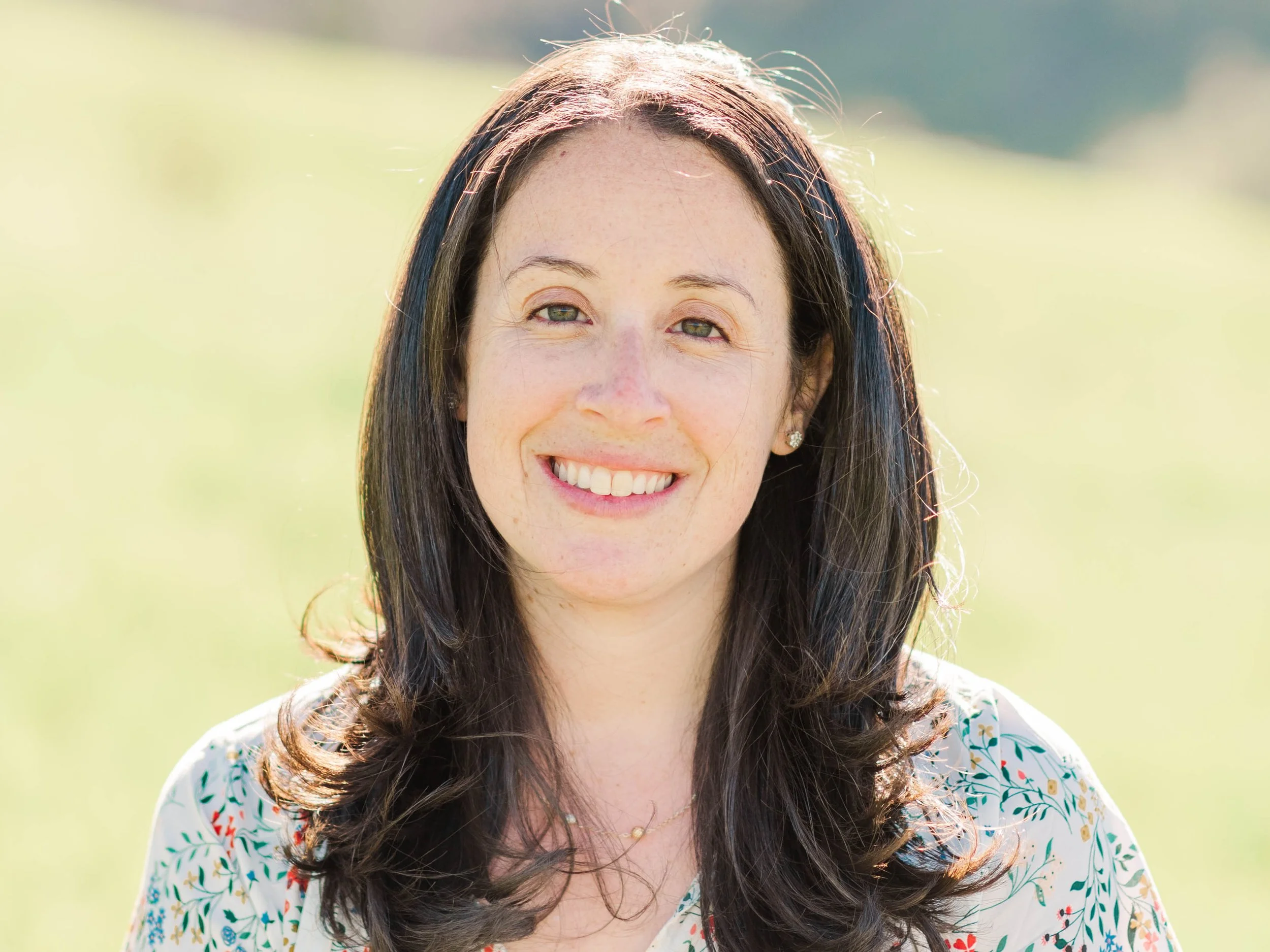 A woman with long dark hair smiling outdoors on a sunny day, wearing a floral patterned top.