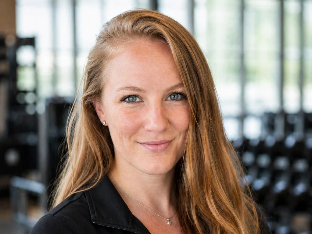 A smiling woman with long red hair and blue eyes, wearing a black top and a necklace, in a gym with equipment and large windows in the background.