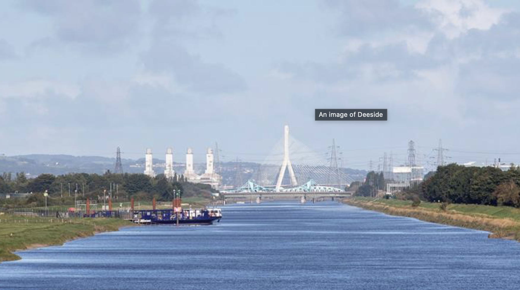 A river with boats docked on the side and a bridge in the distance in Deeside, with industrial chimneys and transmission towers in the background under a partly cloudy sky.