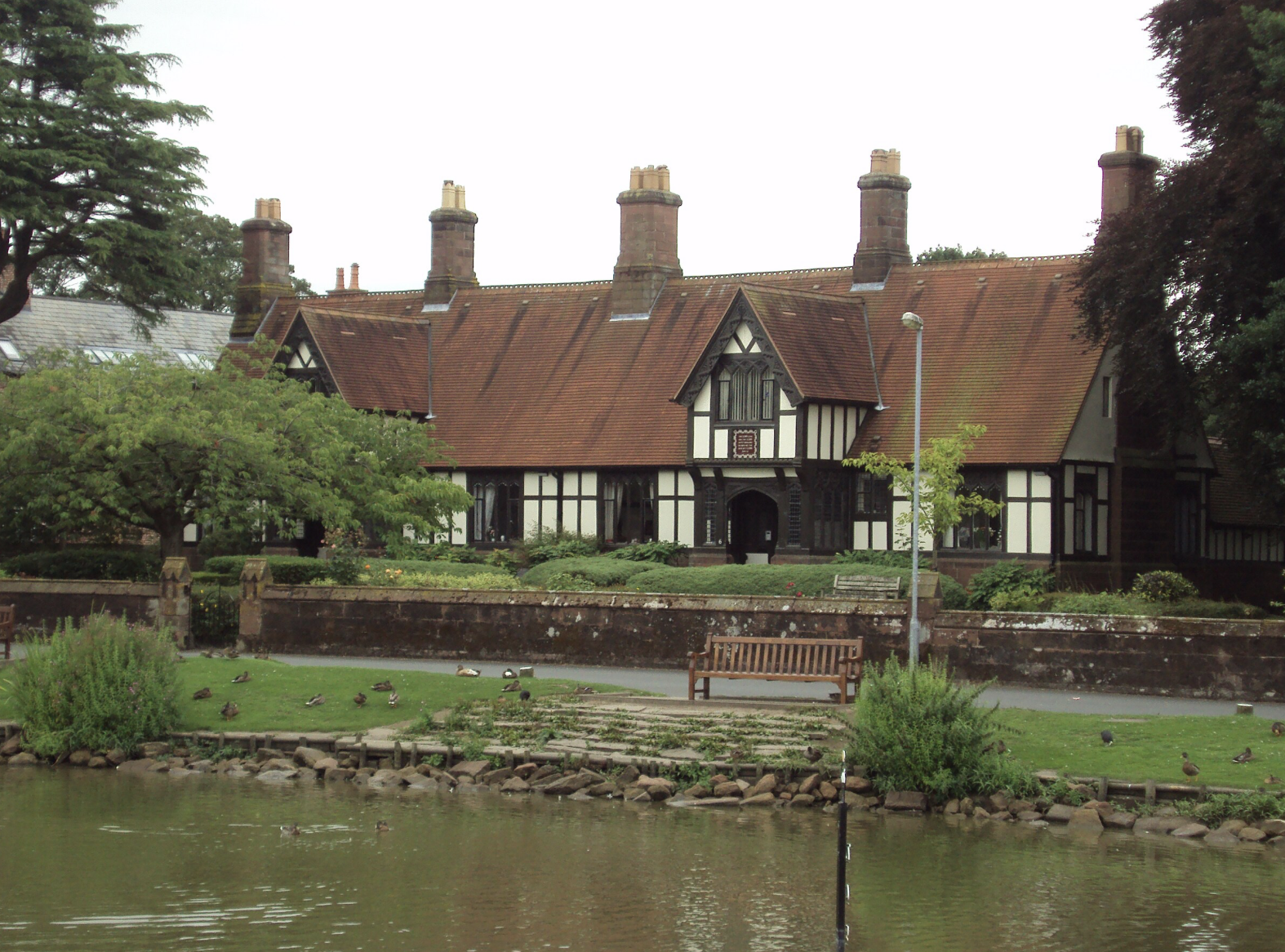 A large Tudor-style house in Christleton with a red-tiled roof, surrounded by trees and a landscaped garden, viewed across a pond with ducks.