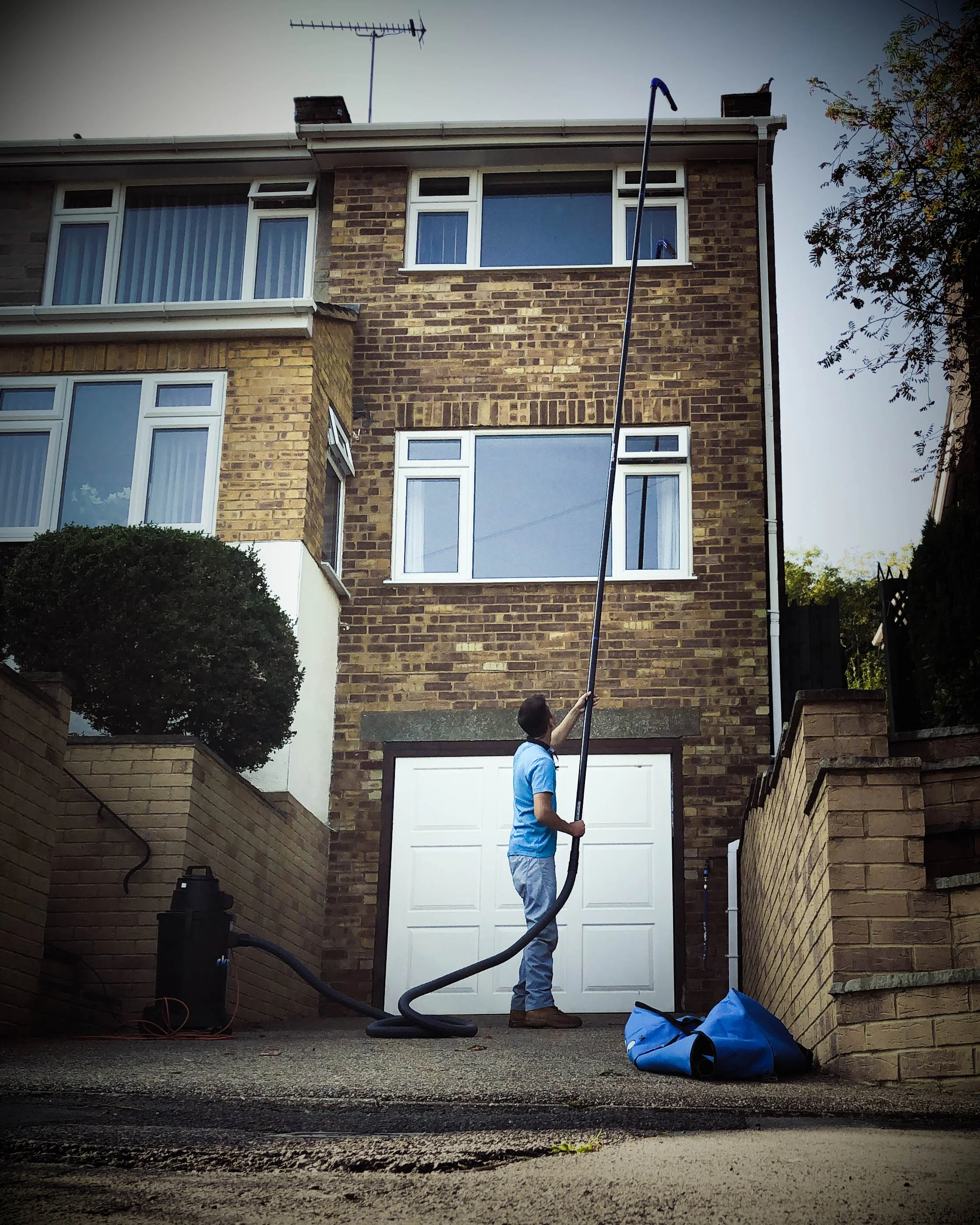 A man in a blue shirt and gray pants directs a vacuum hose spray at a tall window on a brick house. The house has several floors and multiple windows, with a garage door at the base. There is a blue bag on the ground nearby.