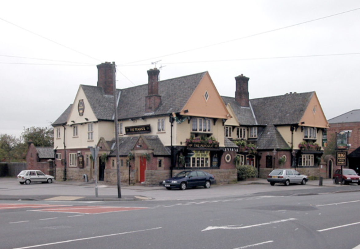 A large pub building with multiple chimneys, beige and pink exterior walls, and decorative flower boxes on the windows. Cars are parked in front and a street with marked lanes is visible. The pub's sign reads "The Plough" and another sign on the corner says "Big".