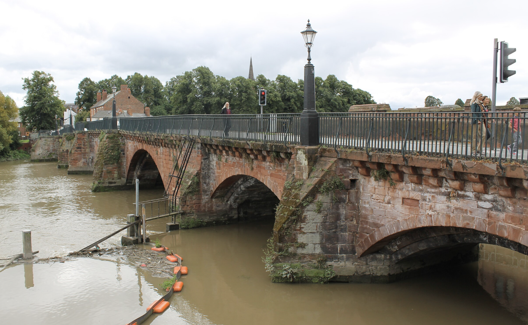 A historic brick bridge in Handbridge, Chester  over a river with pedestrians walking across, surrounded by trees and buildings, overcast sky.