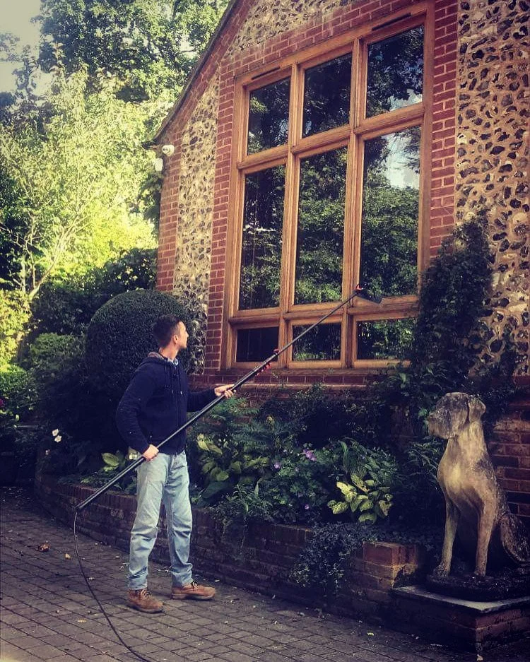A man using a pressure washer to clean a large window on a brick and stone house. There is a garden with bushes and flowers beside him, and a dog sculpture nearby.