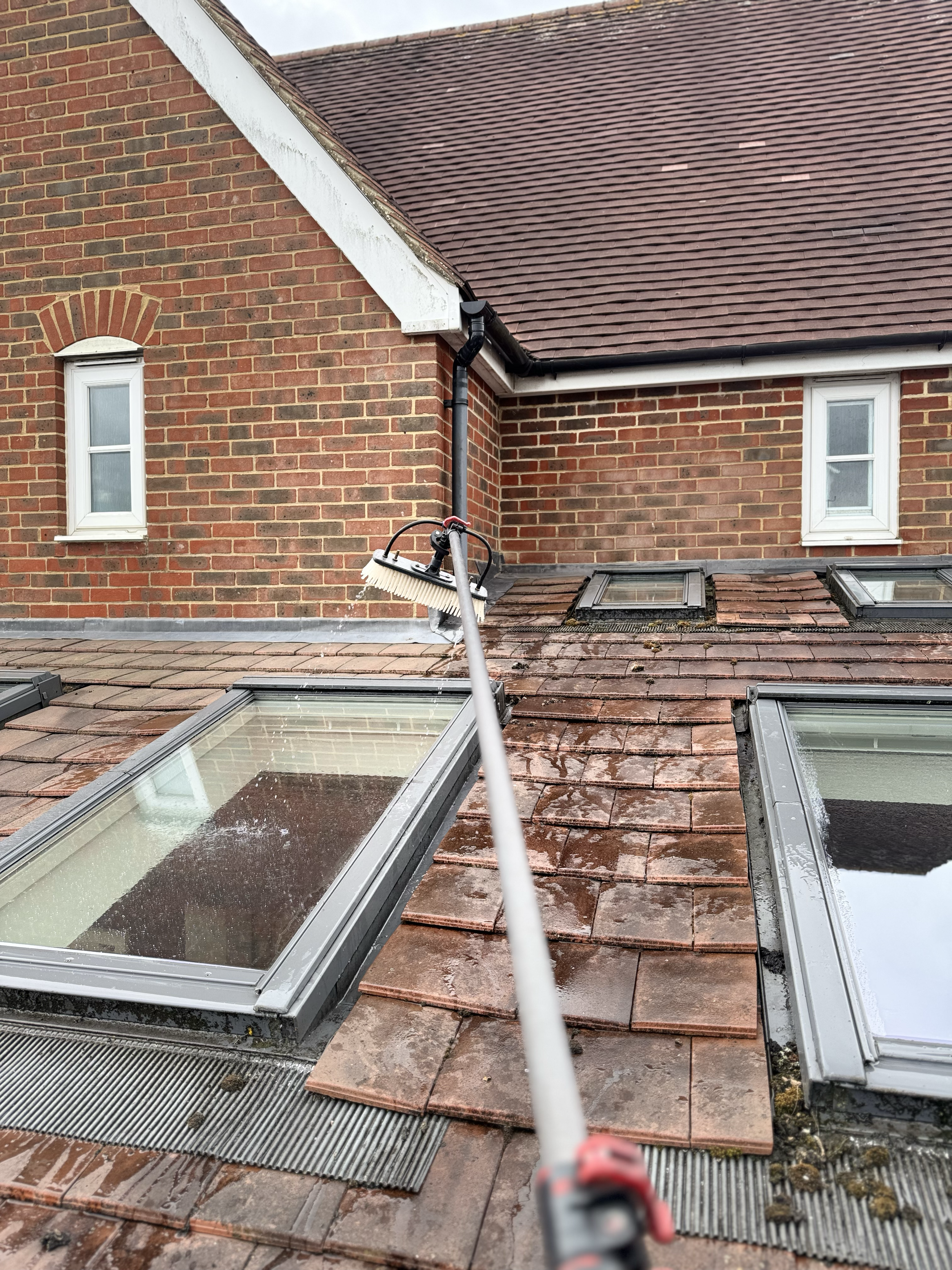 A person cleaning a rooftop with a brush attached to a pole, focusing on skylights and roof tiles on a red brick house.