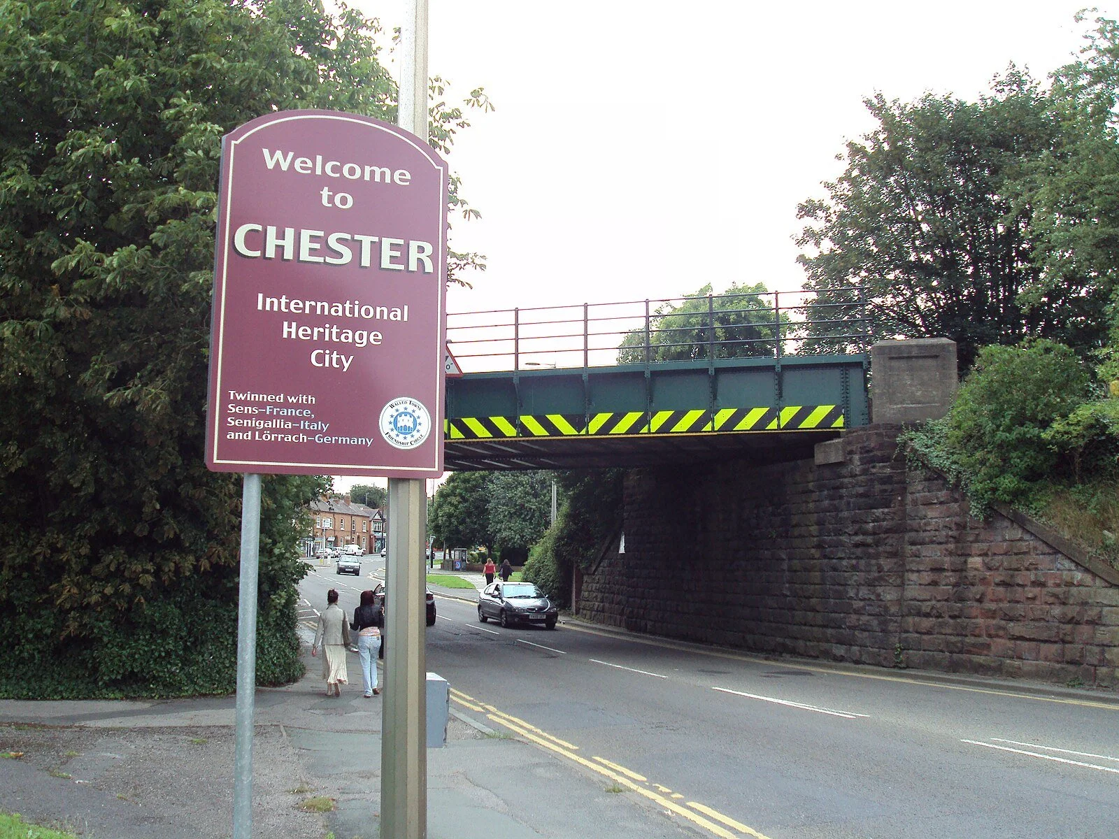 A street view with a purple sign welcoming to Chester, a heritage city, mentioning its twin cities in France, Italy, and Germany. There are pedestrians walking and cars on the road, with a bridge overhead and trees in the background.