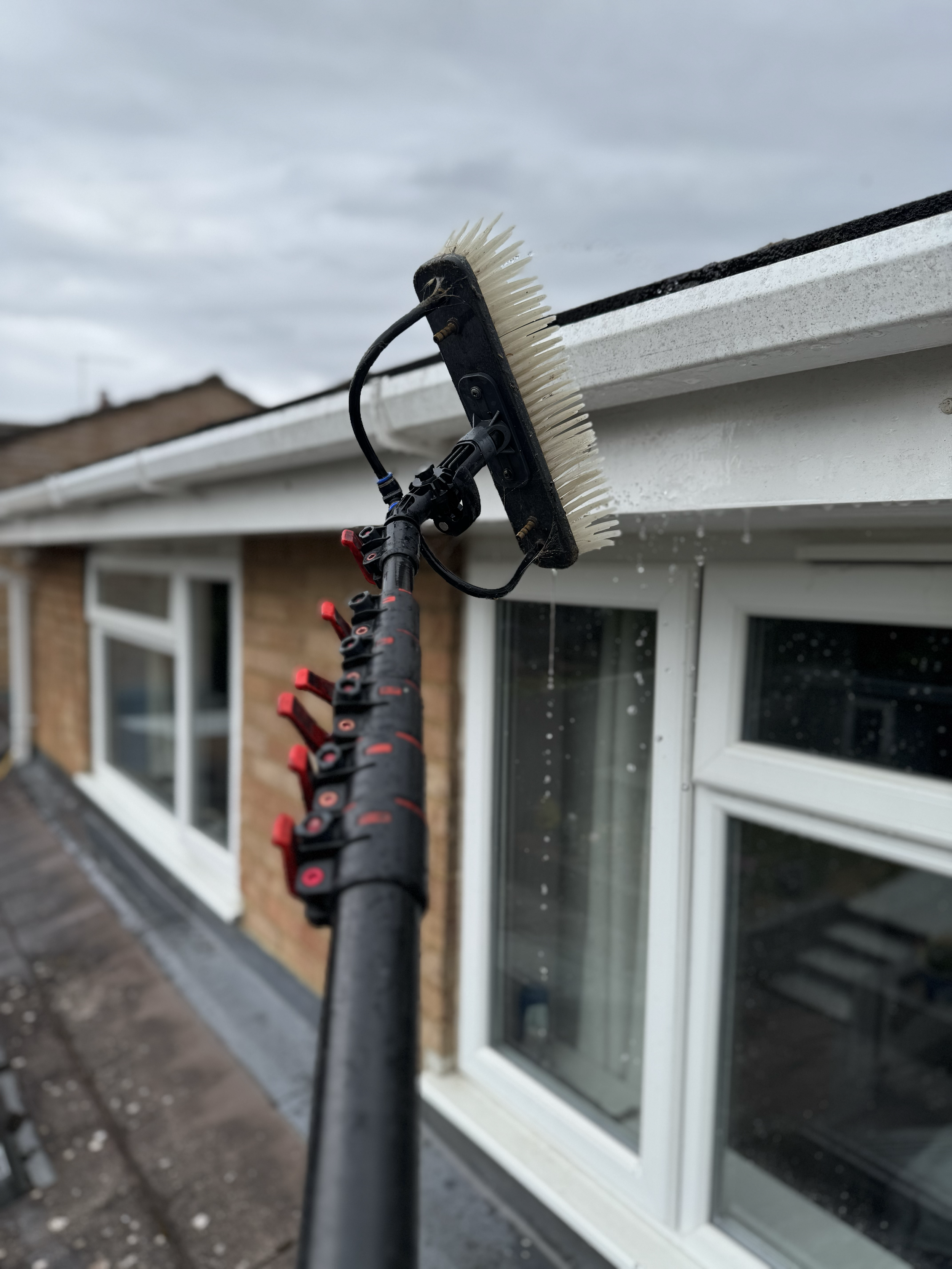 A person cleaning the roof gutter with a specialized tool that has a brush and extends from a scaffold, against a cloudy sky.