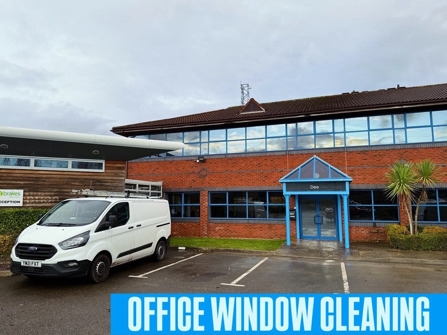 Parking lot in front of a modern office building with blue window frames and a red brick facade, a white van parked, and a sign that reads 'OFFICE WINDOW CLEANING' at the bottom.