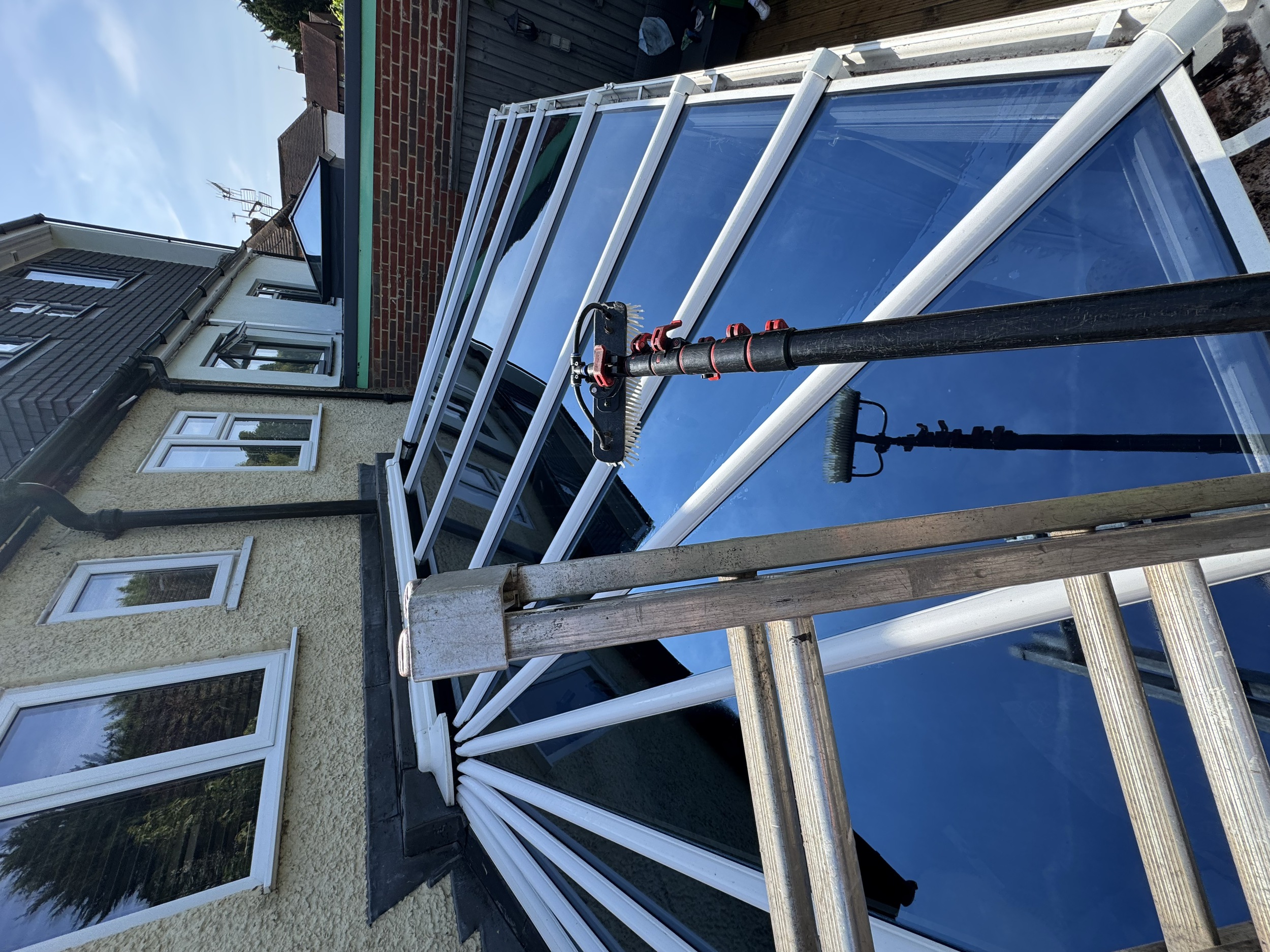 A ladder leaning against a house beside a glass roof extension, with a reflection of the house and sky in the glass.