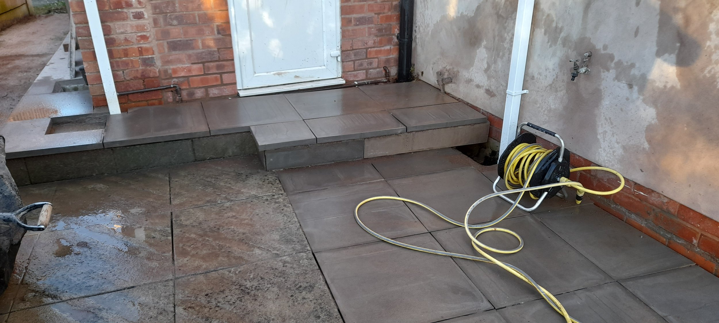 A small set of newly installed grey tiles and bricks creating a step outside a house. A yellow extension cord is coiled on a reel, resting on the tiles."}