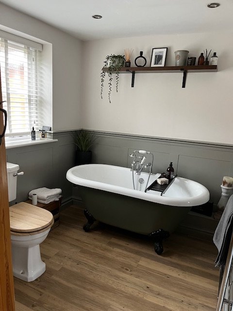 Modern bathroom with a glass-enclosed shower, a white toilet, a navy blue vanity with black hardware, and a large mirror with a wooden frame. Decor includes a vase with dried flowers and small wall shelves with plants.