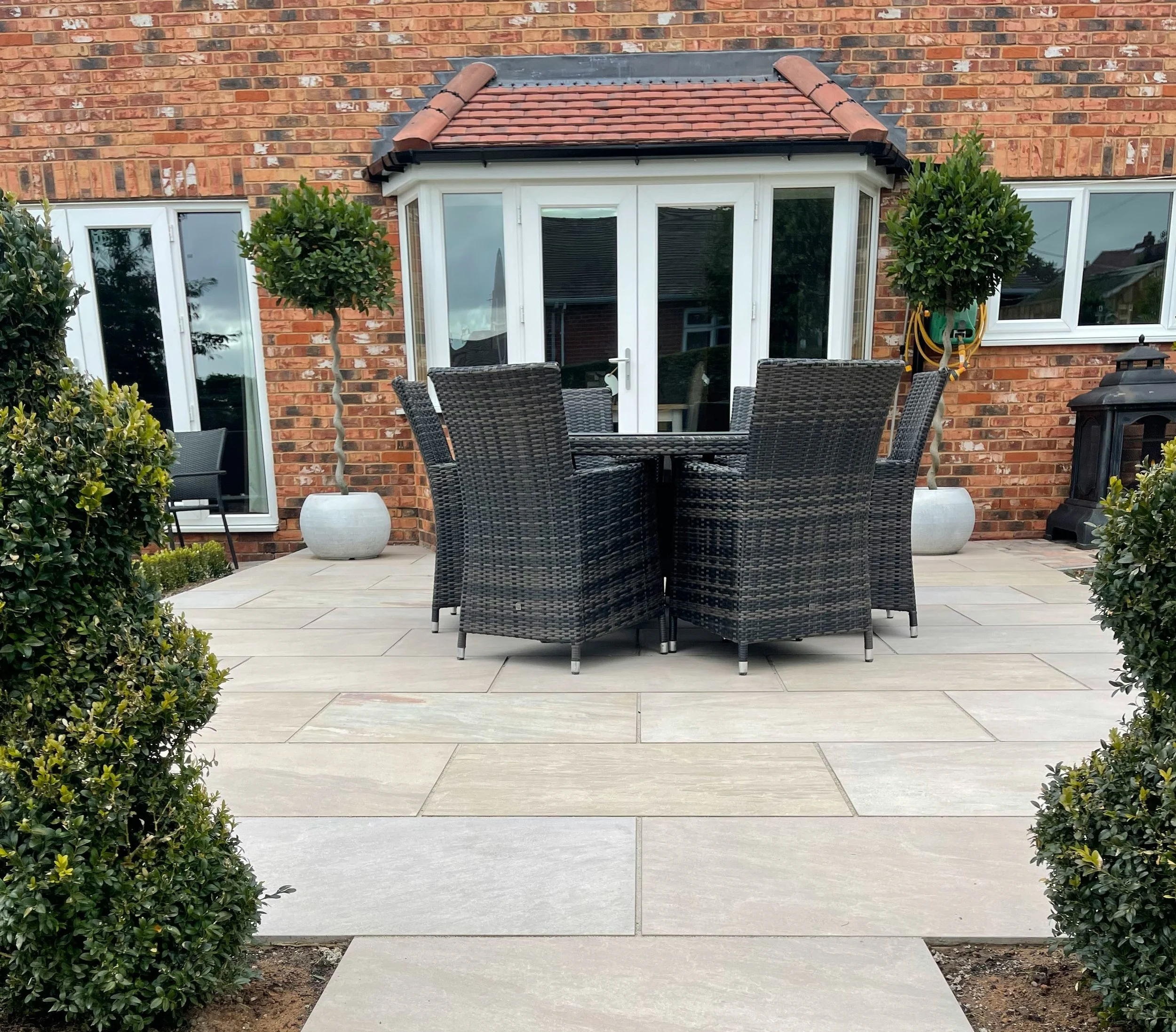 Outdoor patio area with a round black wicker table and six matching chairs on tiled surface, flanked by two trimmed bushes with potted trees behind, and a brick house with white double doors and windows in the background.