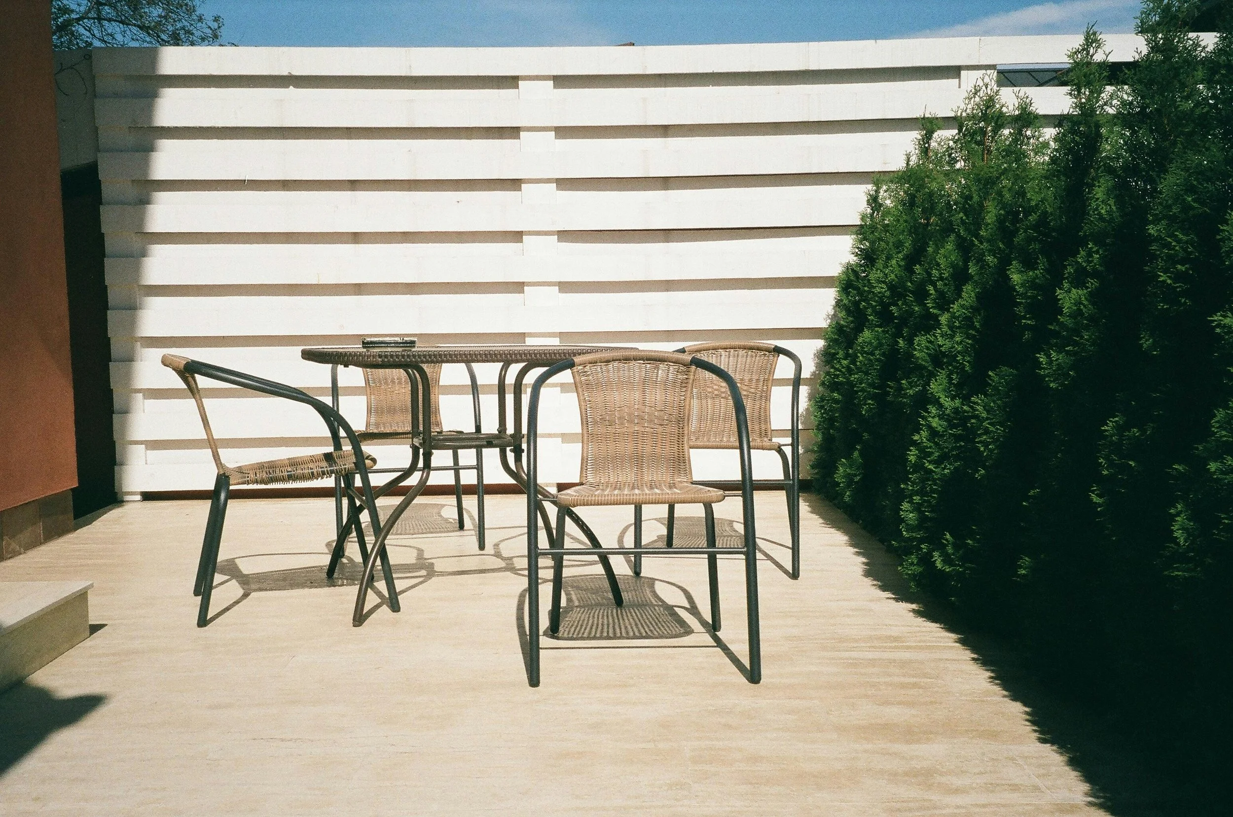 Outdoor patio with four wicker chairs and a glass-topped table, surrounded by a white horizontal slat wall and green bush.