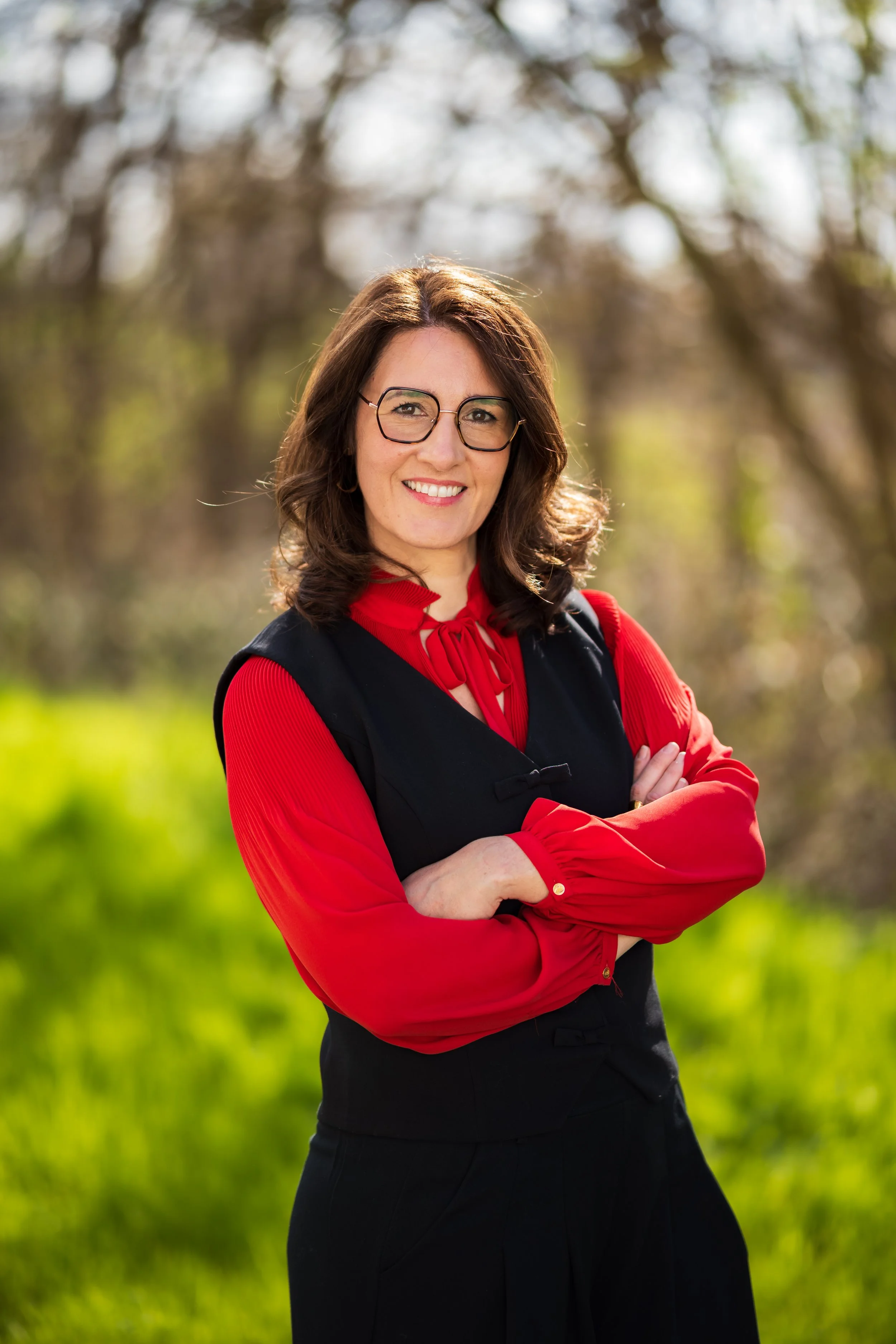 Une femme souriante avec des lunettes, portant un haut rouge et un manteau noir, posant dans un parc avec des arbres en arrière-plan, ensoleillé.