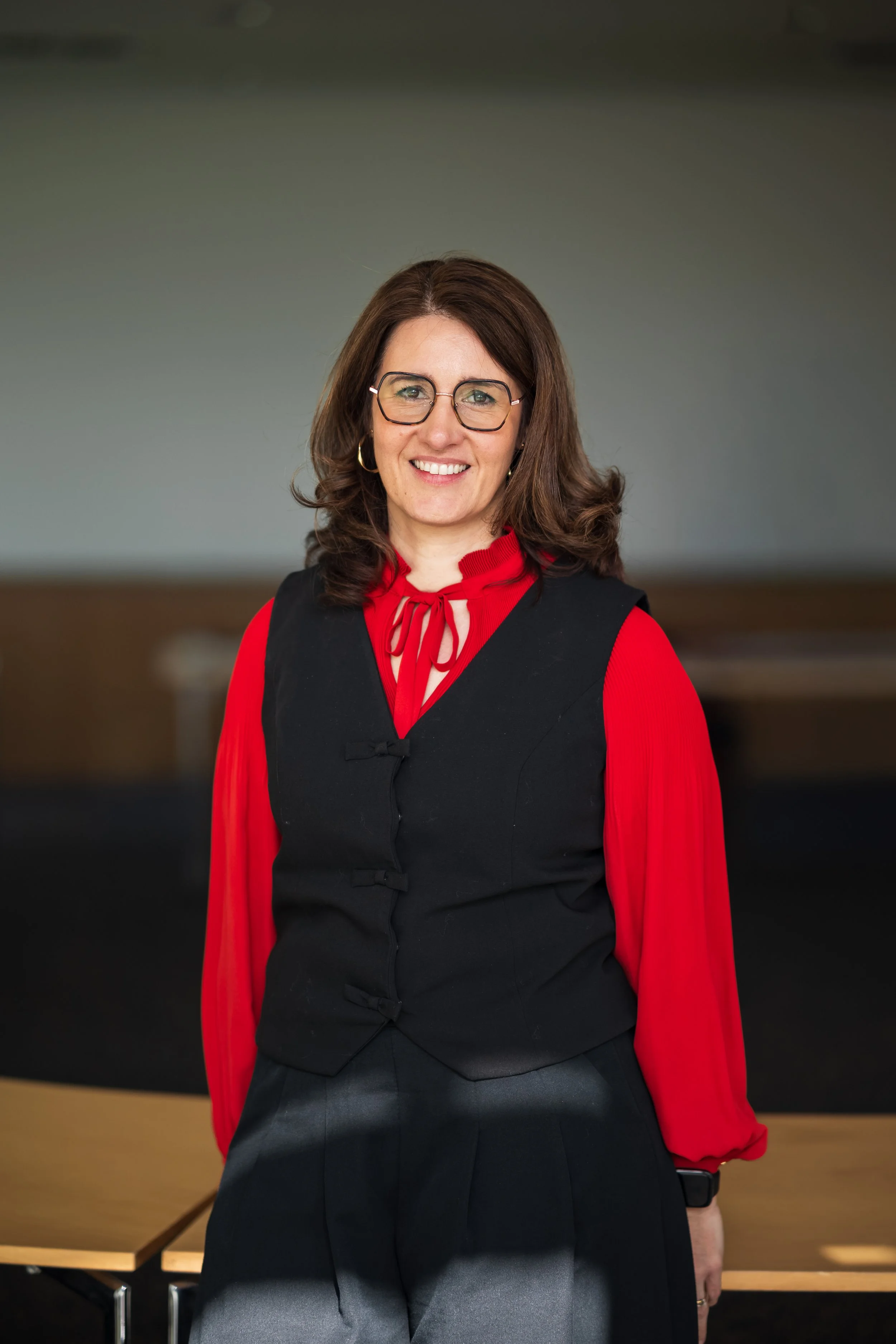 Une femme souriante portant des lunettes, un haut rouge avec un col en ruban, un pull noir et une jupe gris foncé, posant à l'intérieur d'une pièce.