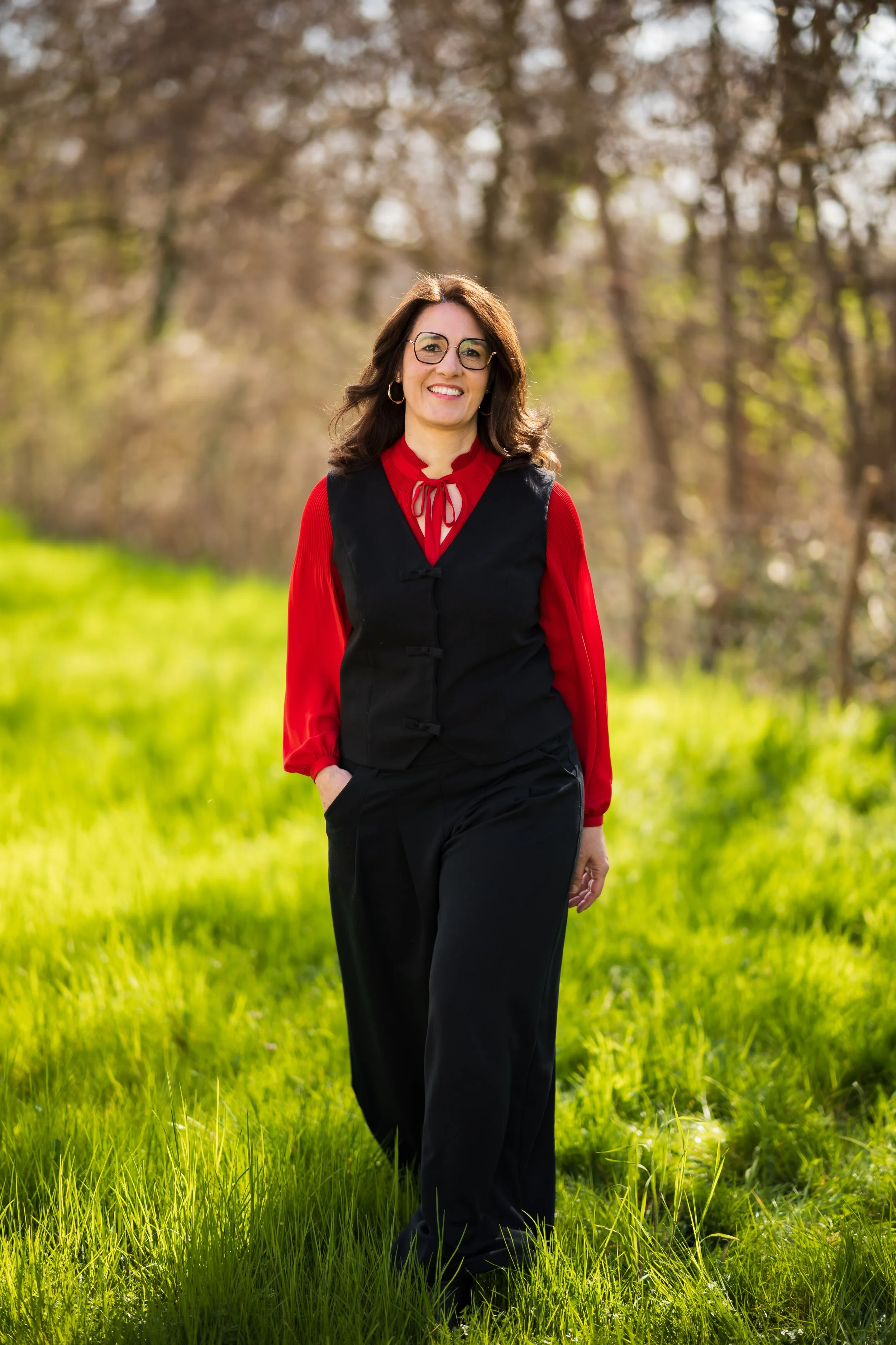 une femme souriante portant une veste noire et une chemise rouge avec un nœud en haut, debout dans un champ vert avec des arbres en arrière-plan, en pleine journée ensoleillée