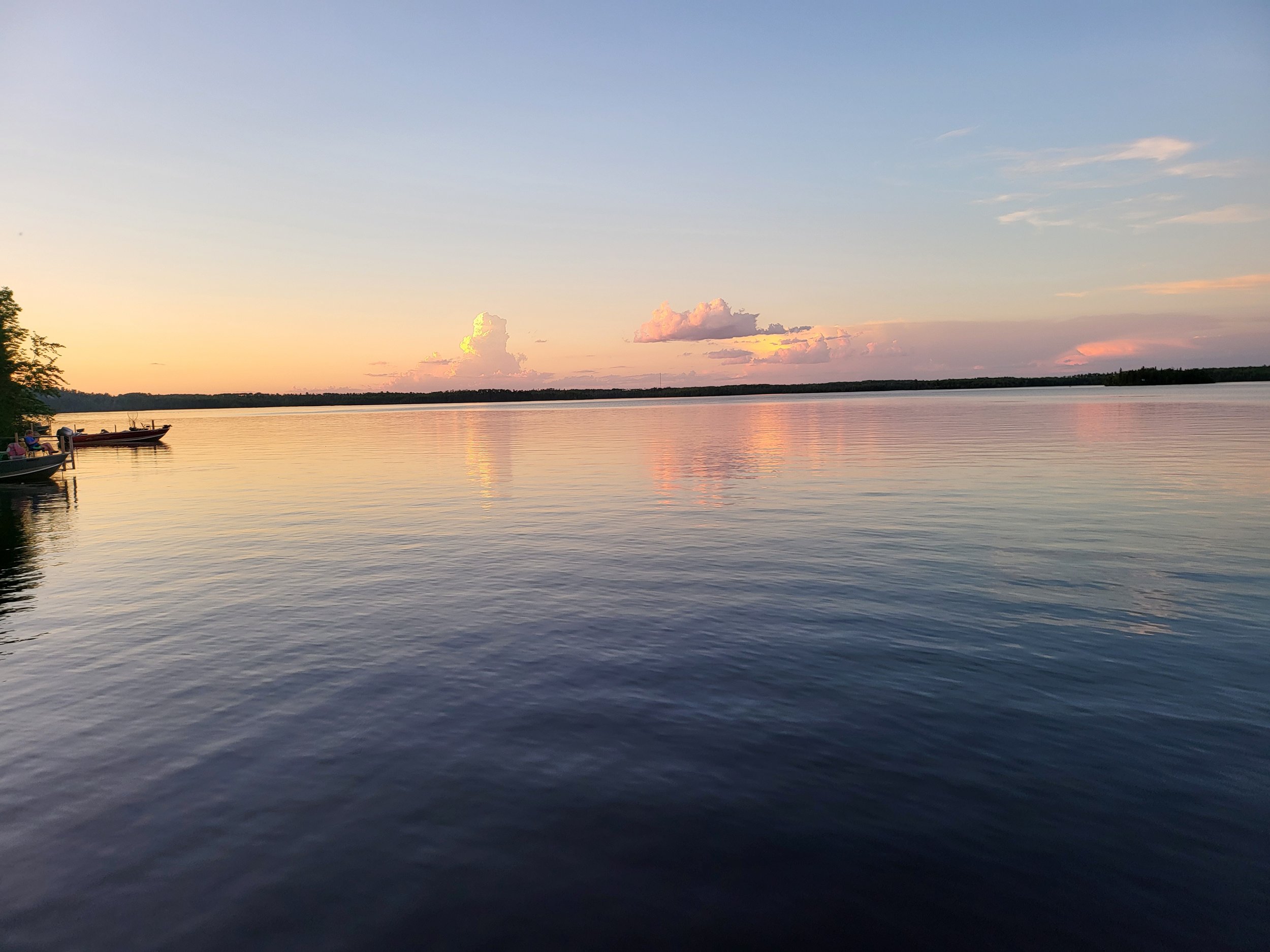 Serene lake at sunset with reflection of colorful clouds on water, two boats on left, and distant tree-lined horizon.