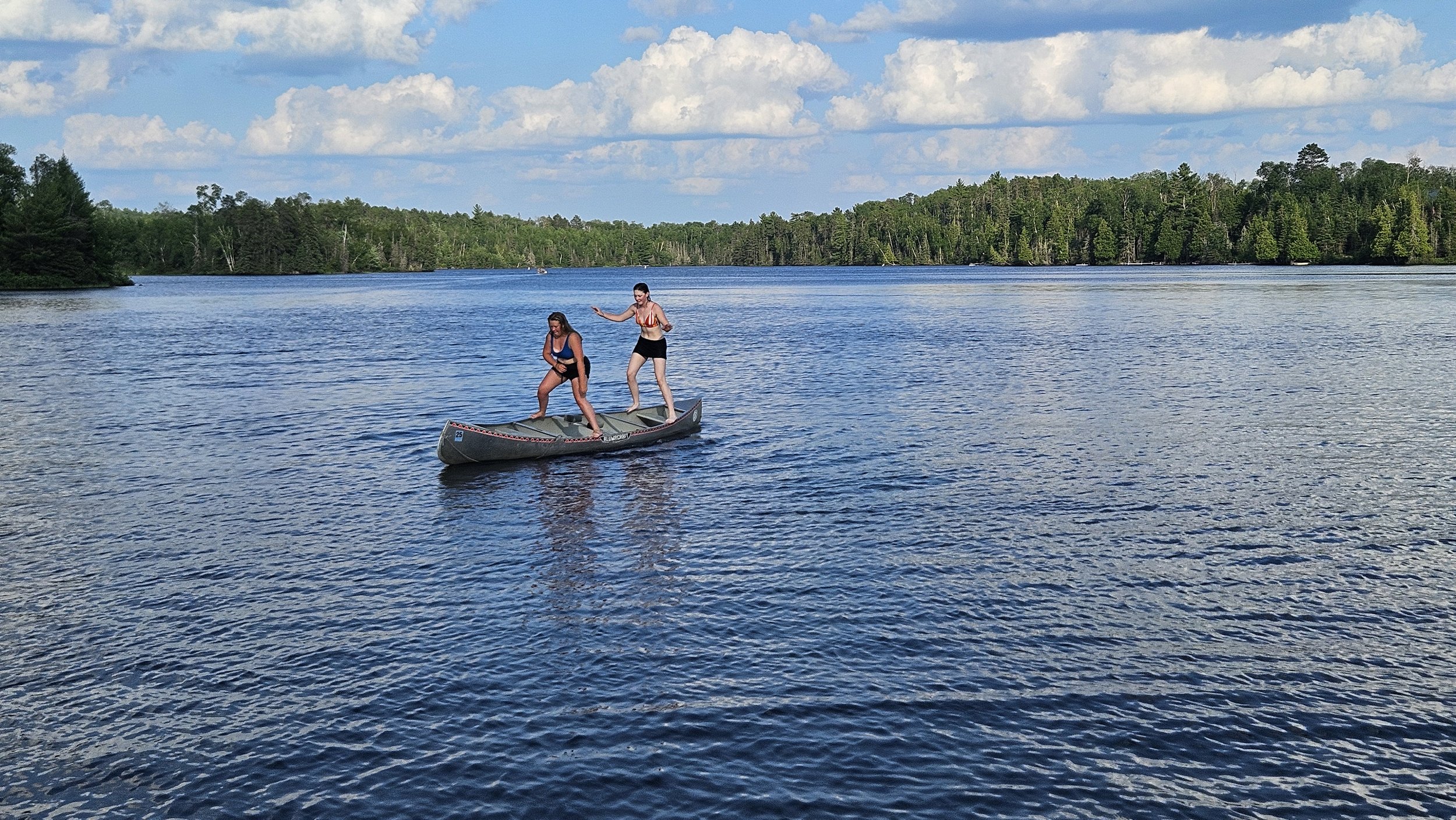 Two people standing on a canoe in the middle of a lake, surrounded by trees and a cloudy sky.