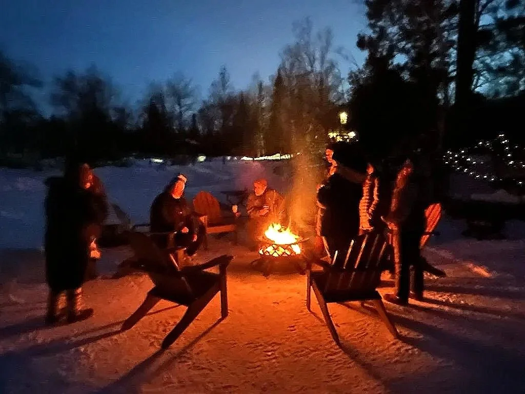 Group of people sitting and standing around a campfire in a snowy outdoor setting at dusk, with trees in the background and chairs arranged around the fire.