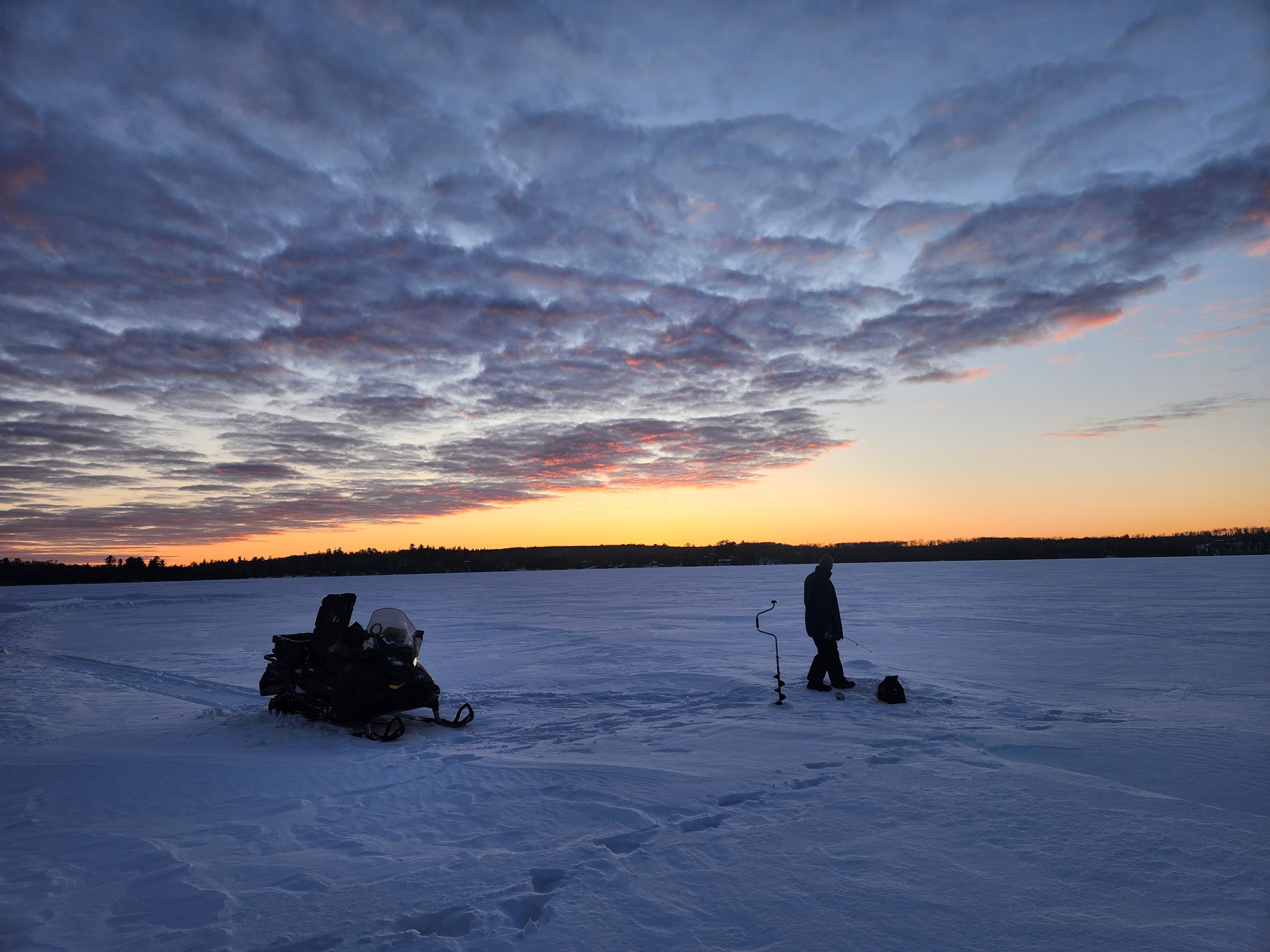 Person ice fishing on a frozen lake at sunset with a snowmobile nearby.
