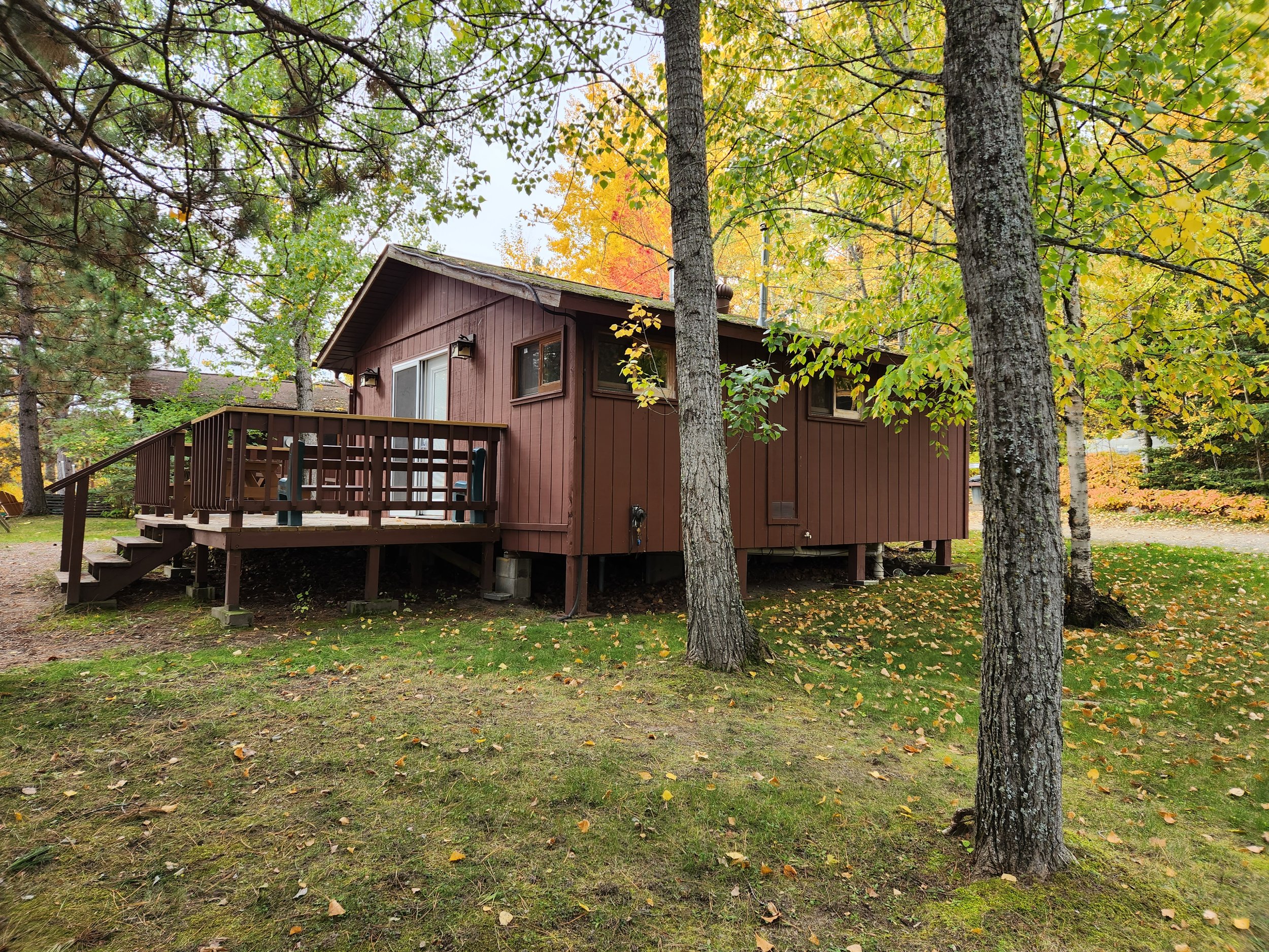 A small brown cabin with a wooden deck and stairs, surrounded by trees with green and yellow leaves, in an autumnal forest setting.