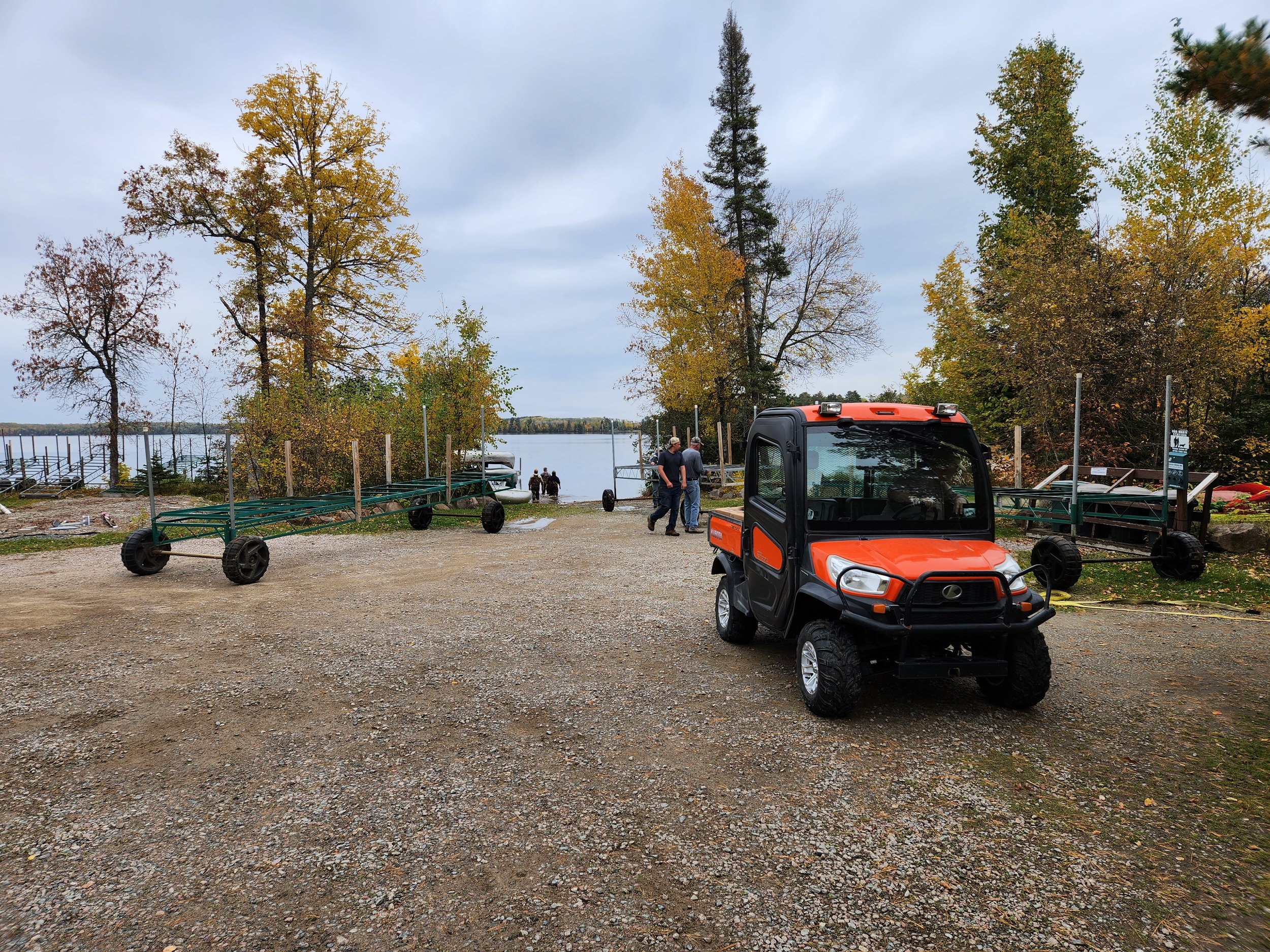 A red utility vehicle parked on a gravel path near a lake, surrounded by autumn trees and several trailers. People are standing near the water's edge.