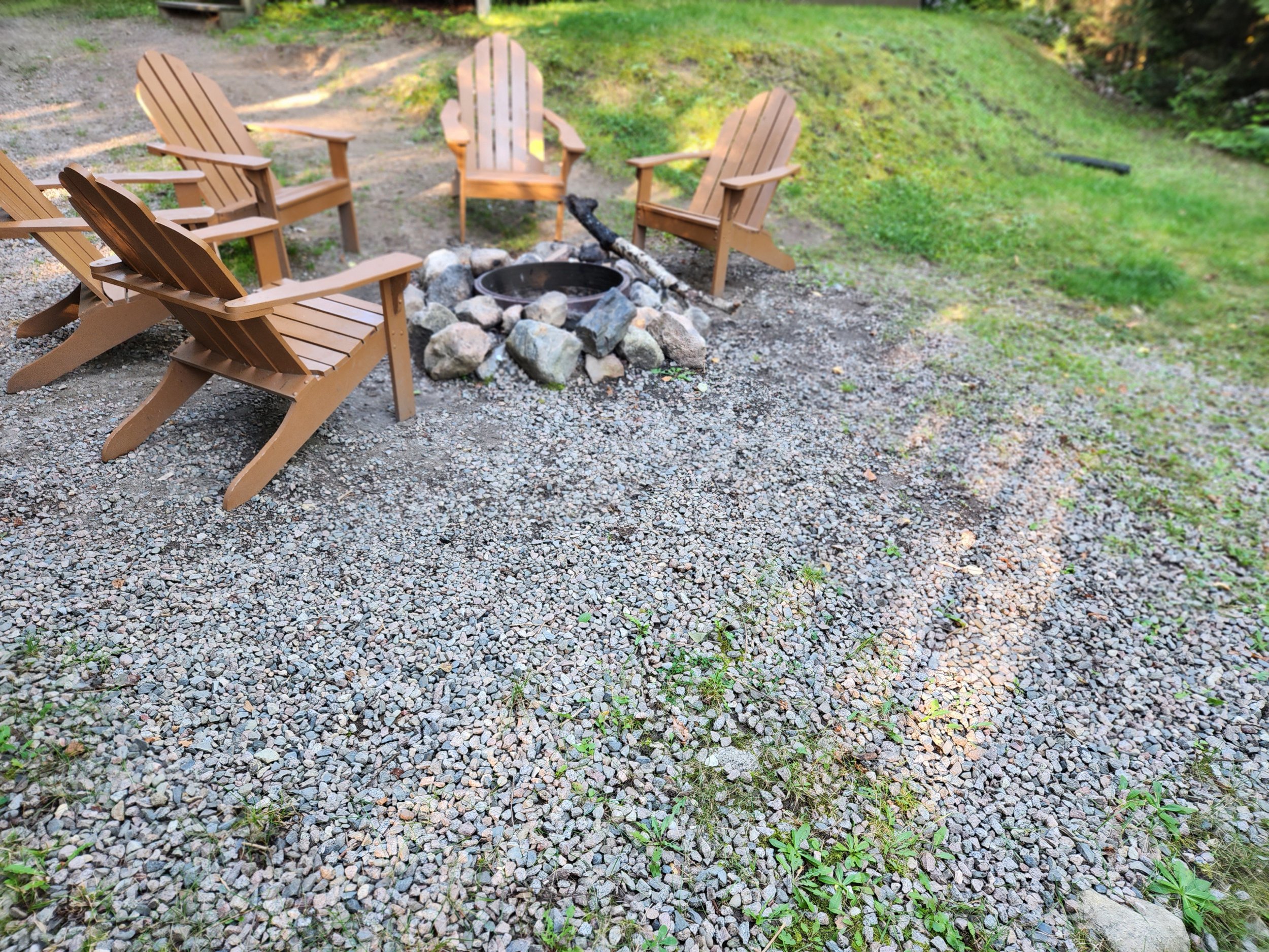 Outdoor fire pit area with four wooden Adirondack chairs surrounding a stone fire pit, set on a gravel surface in a grassy yard.
