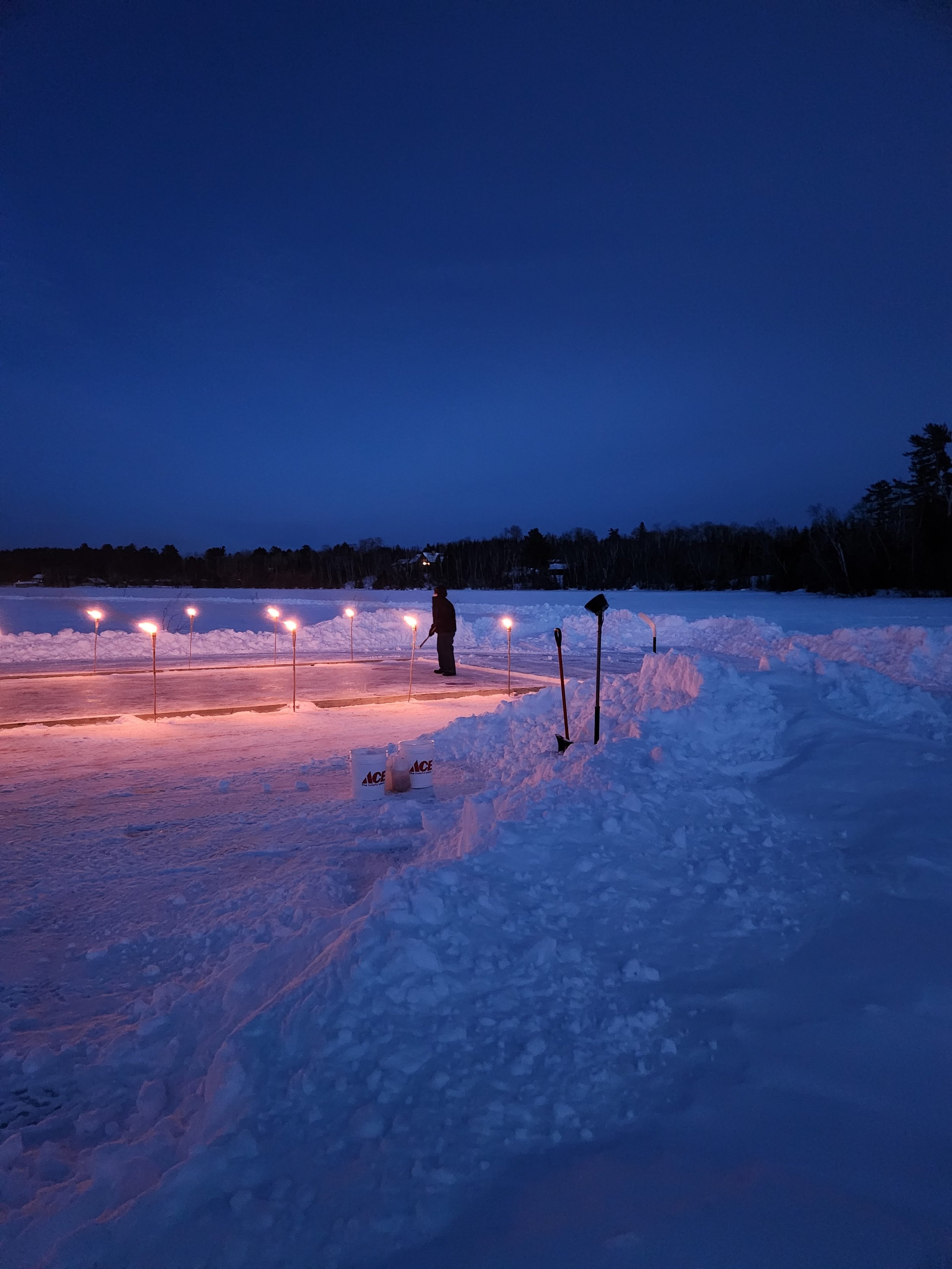 Snow-covered landscape at dusk with a person standing near a lit ice rink; shovels and tools nearby.