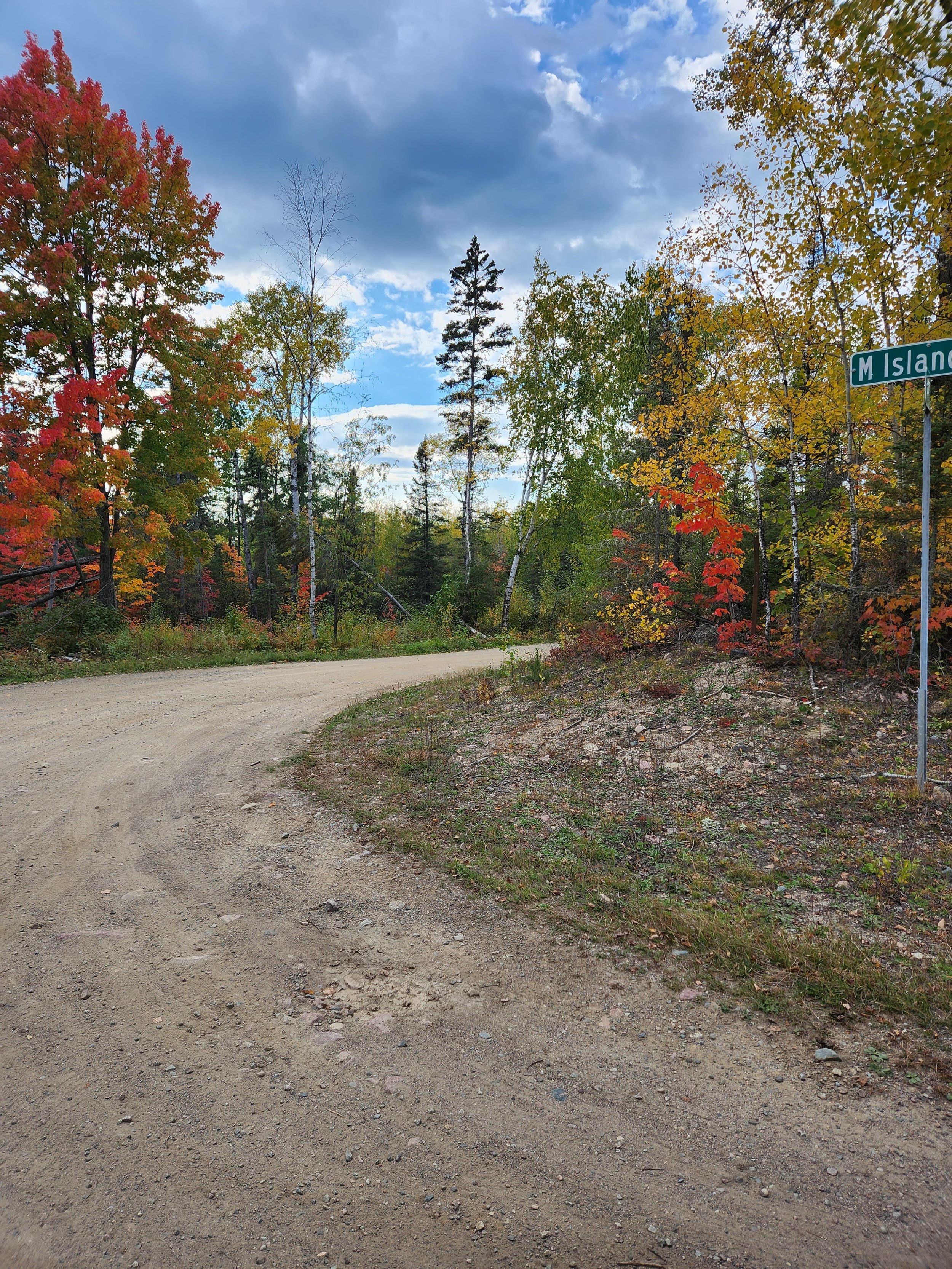 Dirt road curving through a forest with autumn foliage, featuring red, orange, and green leaves. A street sign reads "M Island." Cloudy sky above.