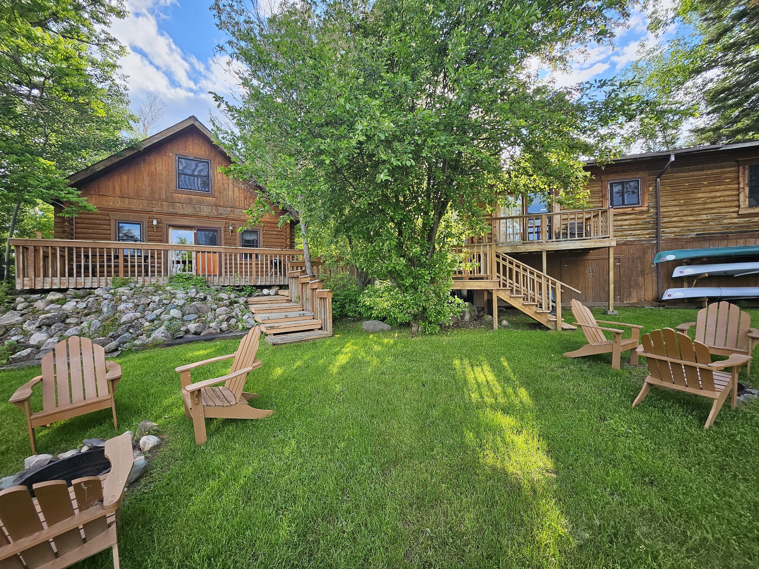 A rustic wooden cabin with a stone foundation, surrounded by lush greenery. Four wooden Adirondack chairs are positioned on a grassy lawn, facing a fire pit. Two sets of stairs lead up to decks on the cabin, with canoes stored under one section. A la