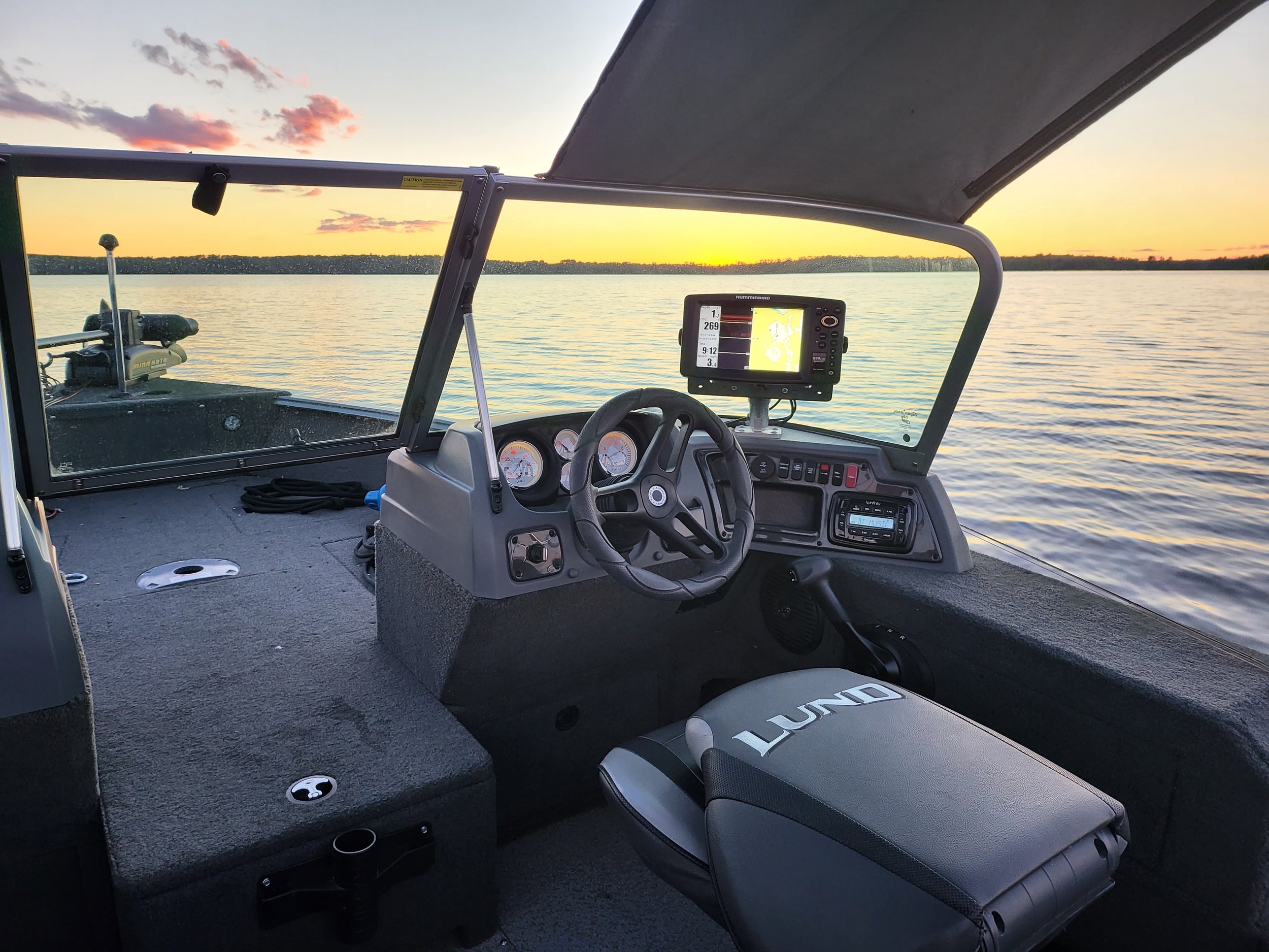 Boat cockpit with steering wheel, instrument panel, and a digital screen displaying navigation. The boat is on a calm lake at sunset, with visible water and a clear sky.