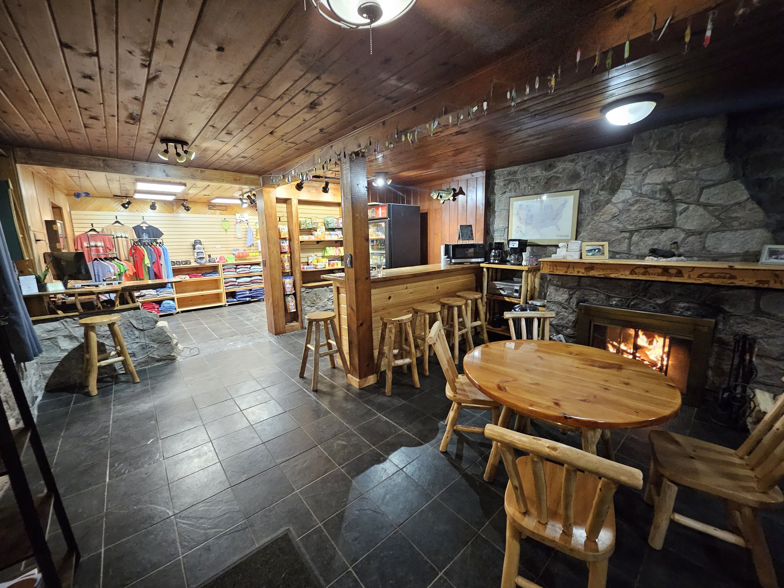 Cozy cabin interior with wooden furniture, stone fireplace, and store section with clothing and snacks.
