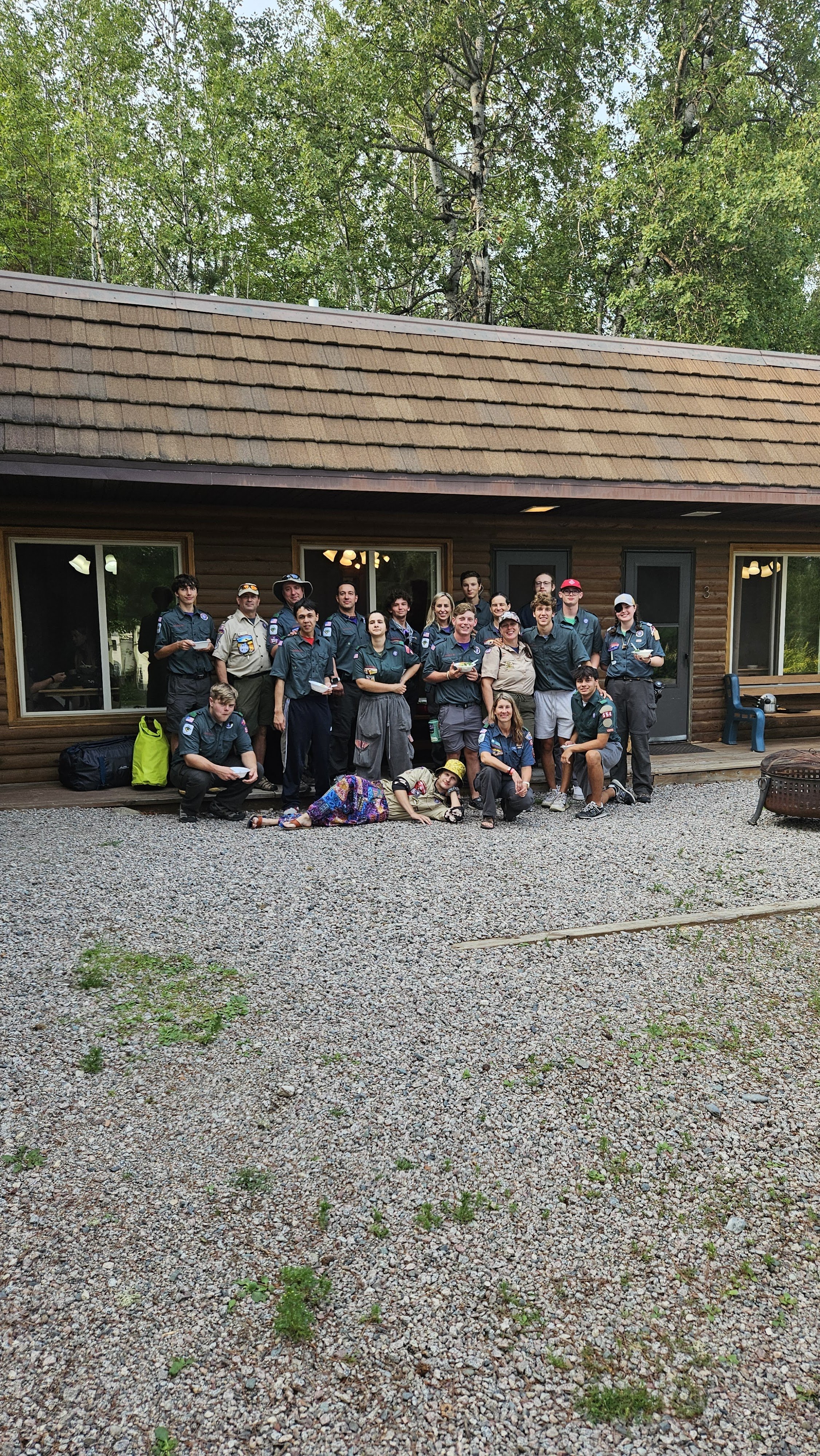 Group of people in matching uniforms standing in front of a cabin with trees in the background.